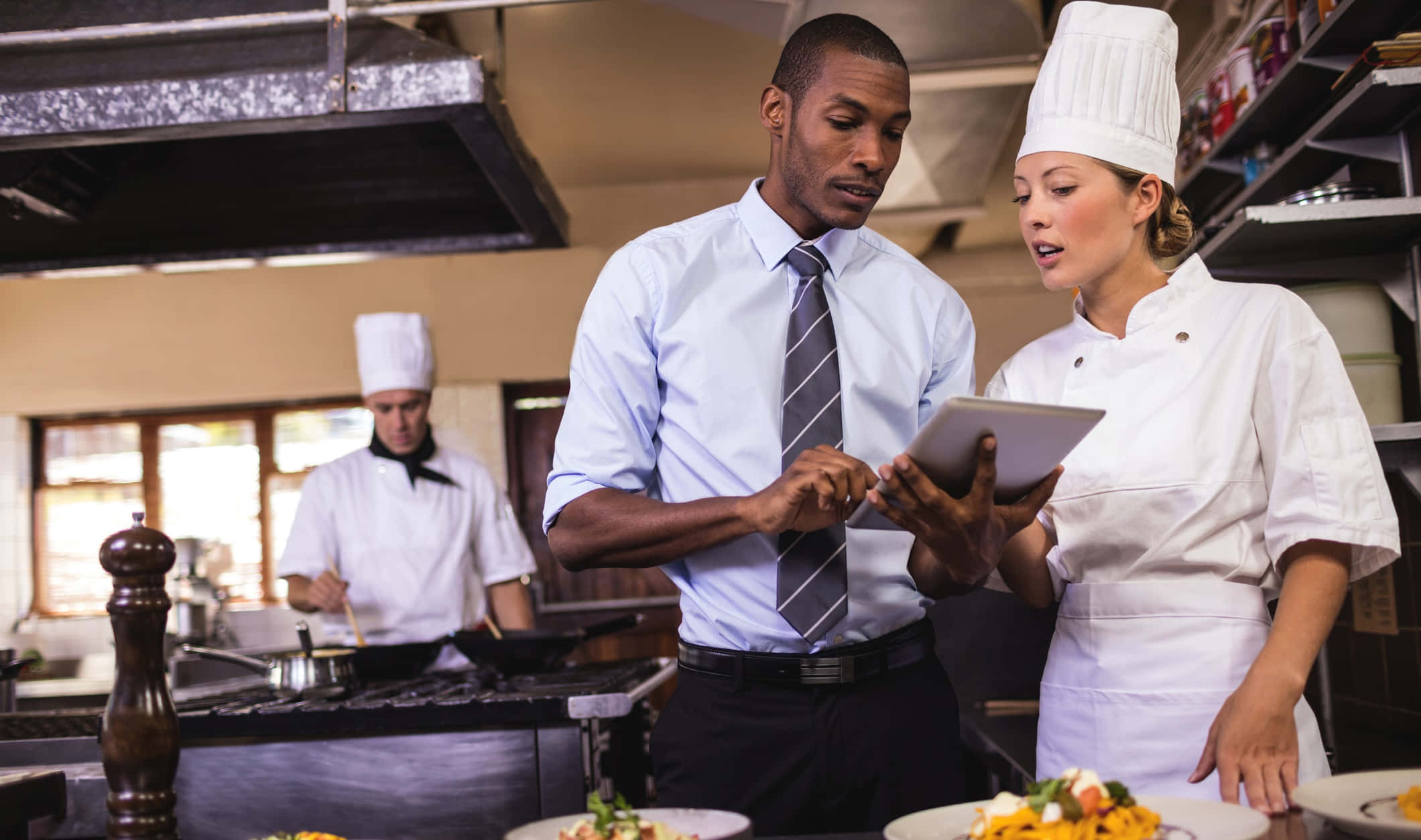 Hospitality 桌布 壁紙 — 一位廚師和經理在繁忙的專業廚房裡查看平板電腦，背景中有擺盤的菜餚和另一位廚師在工作。