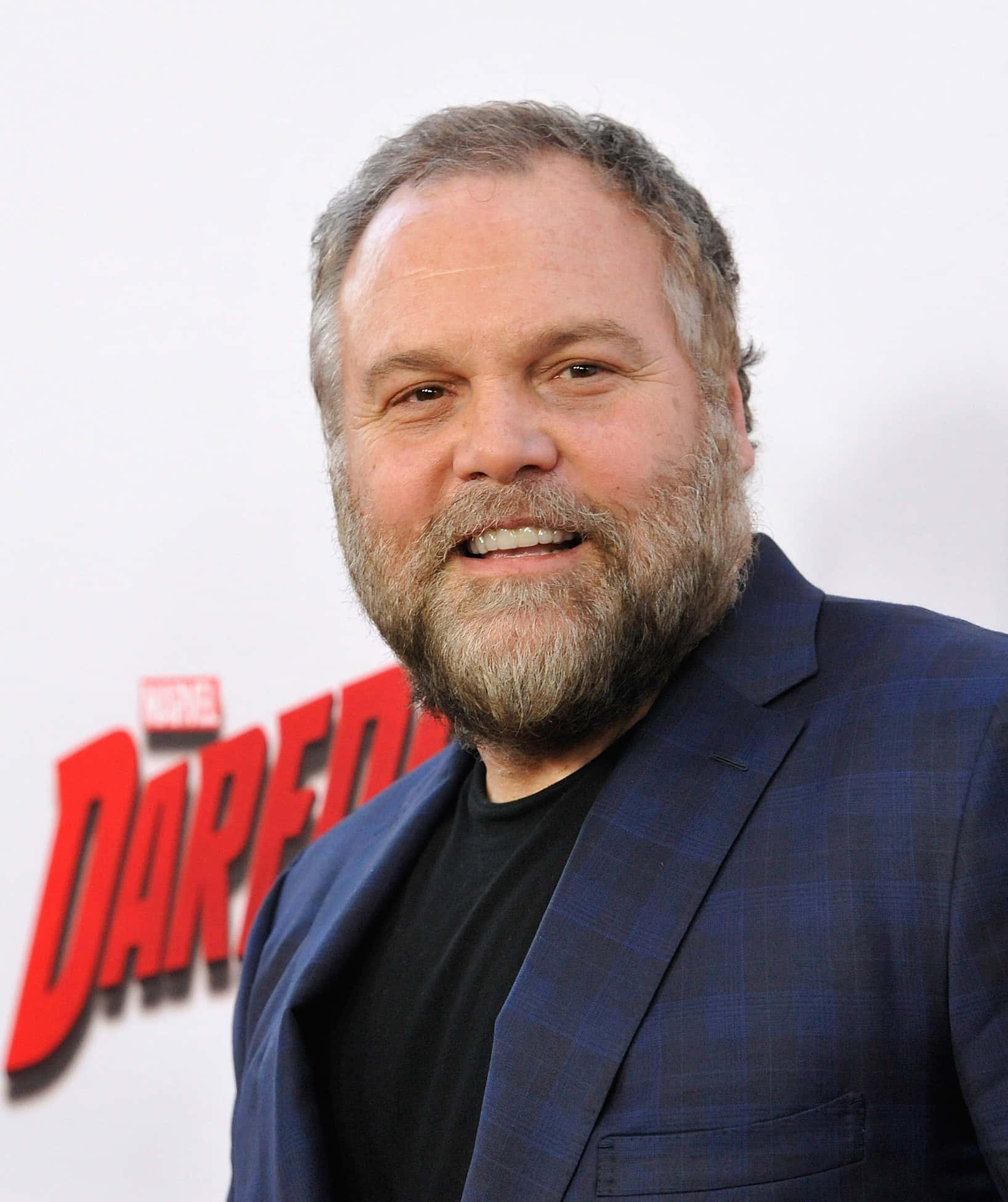 Vincent D'onofrio 桌布 壁紙 — 一位微笑、留著灰白鬍鬚的男子，在白色背景上穿著黑色襯衫外搭藍色格紋西裝外套，並有紅色Marvel Daredevil文字。