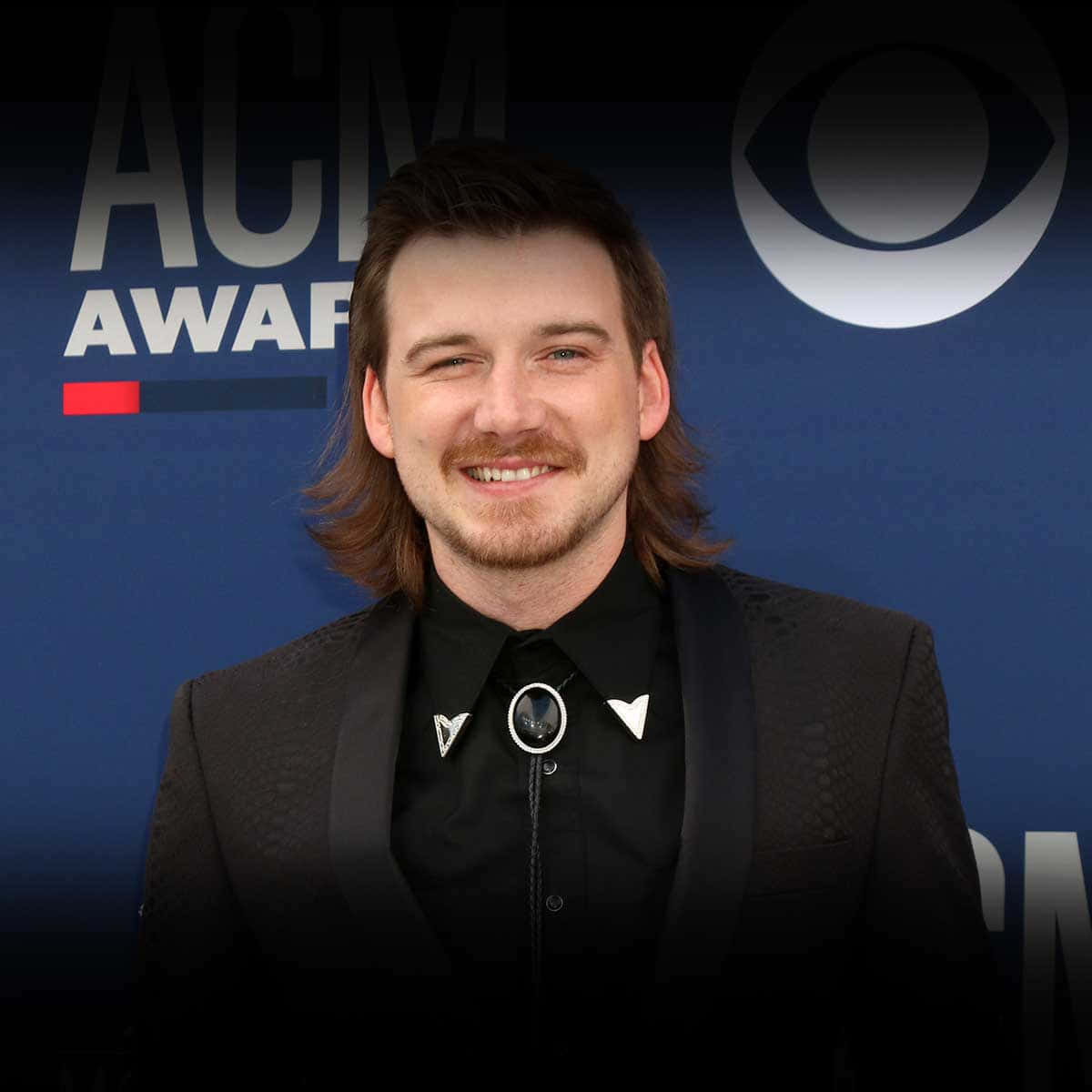 Morgan Wallen 桌布 壁紙 — 一位微笑、長髮、留鬍鬚的男子，身穿黑色西裝和波洛領帶，背景是藍色的ACM Awards。
