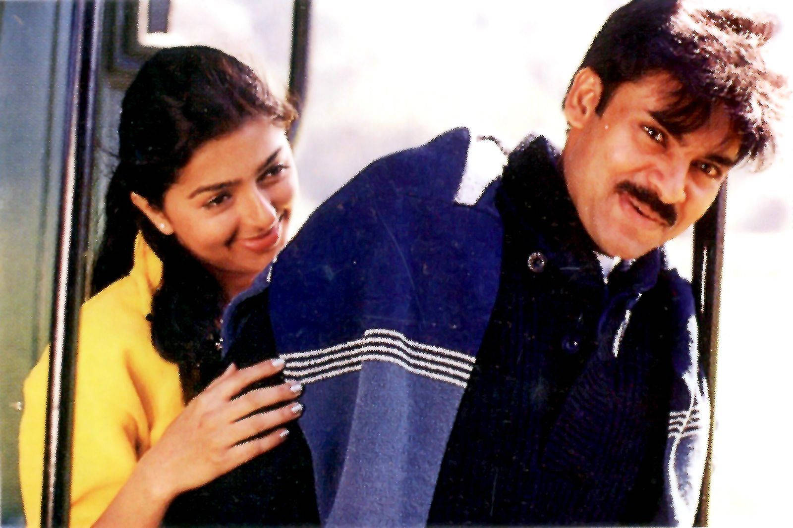 Pawan Kalyan 桌布 壁紙 — 一位微笑嘅女士身穿黃色上衣，深情噉挨住一個著藍色外套嘅男士，企喺門口旁邊。