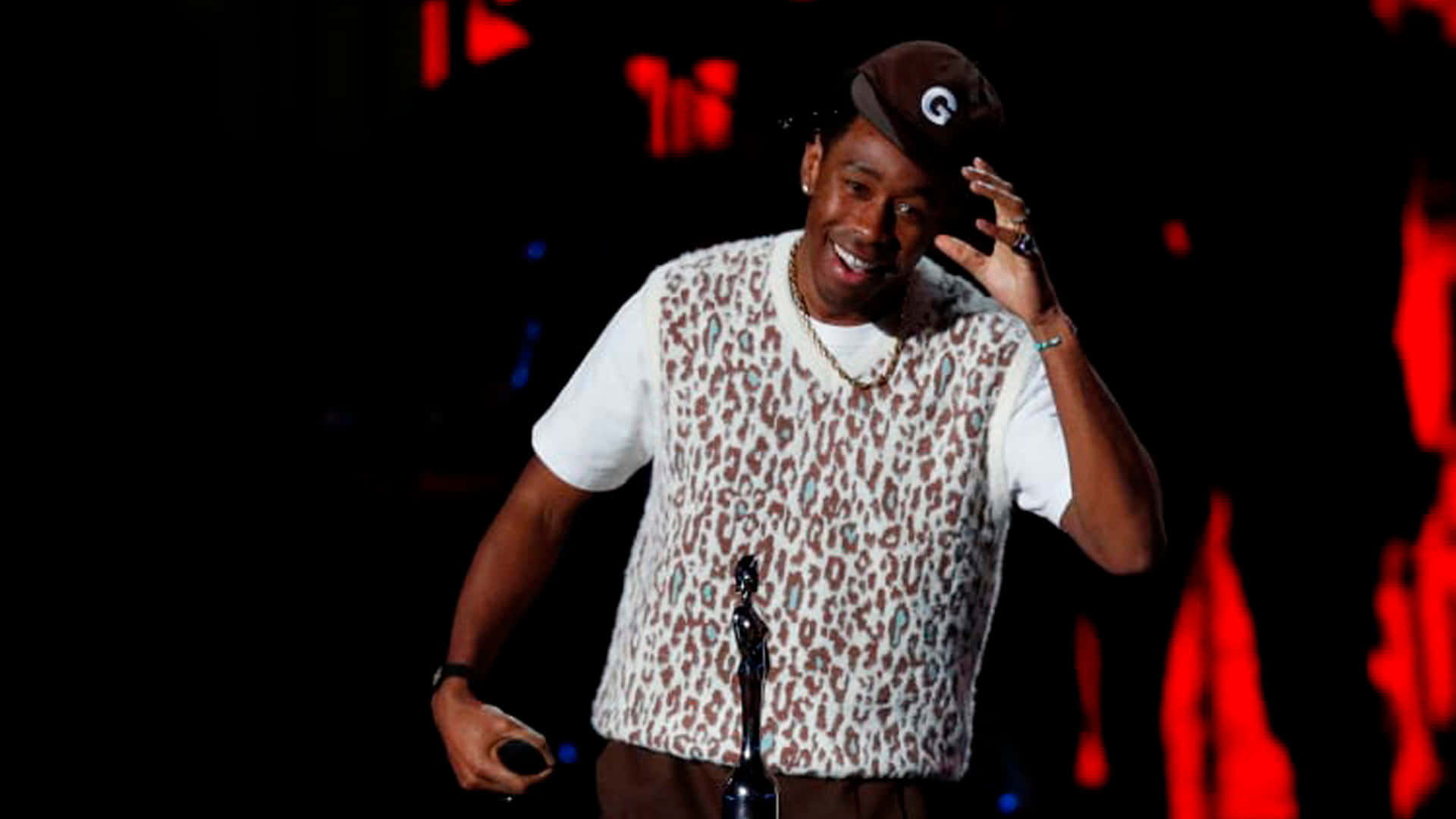 tyler the creator 桌布 壁紙 — 一位微笑嘅男人，身穿豹紋背心同帽，喺紅色燈光嘅黑暗舞台上揮手，同時攞住一個獎項。