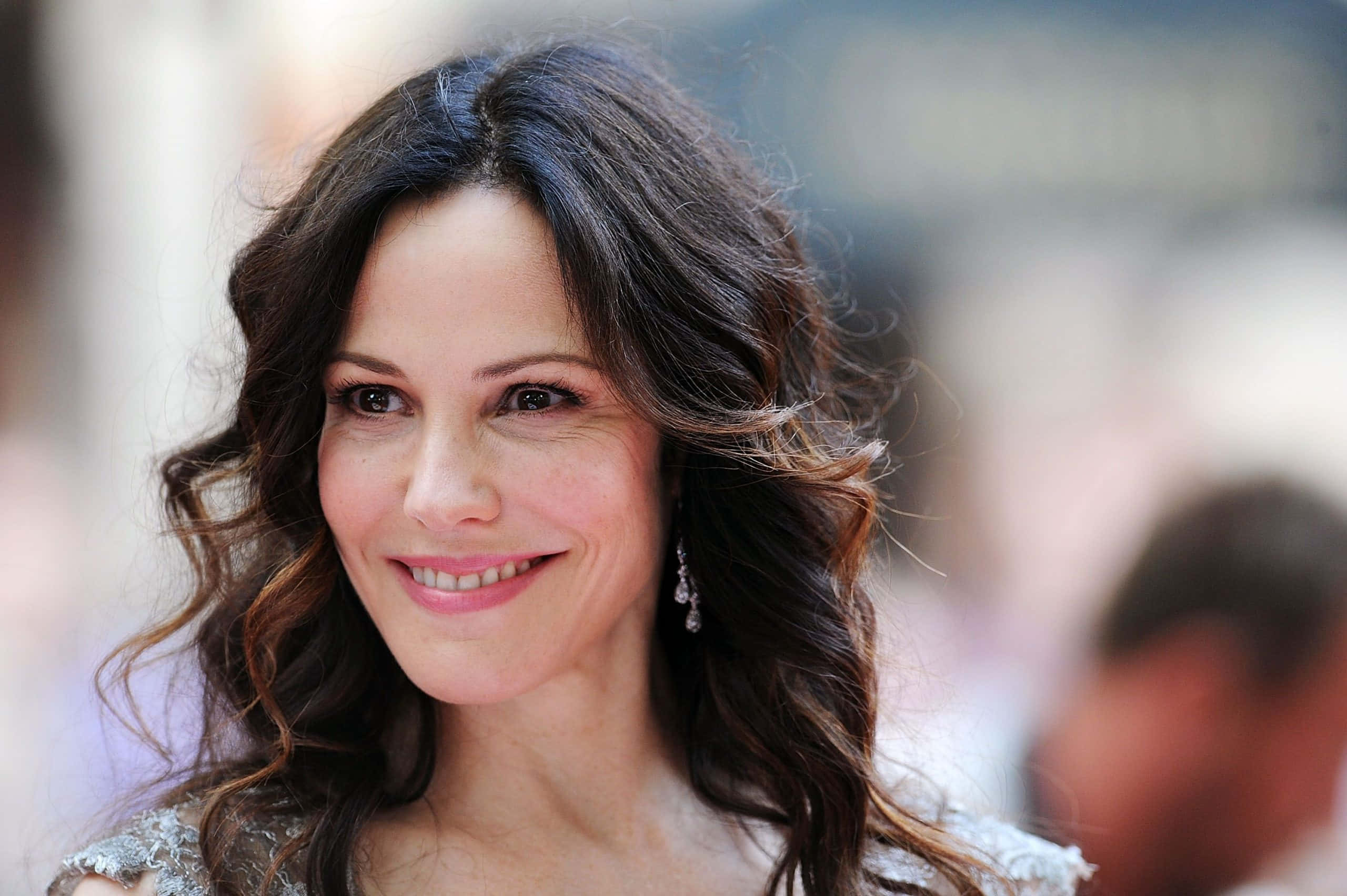 mary louise parker 桌布 壁紙 — 一位微笑女士的 close-up 肖像，擁有波浪深色頭髮和優雅耳環，背景柔和模糊。