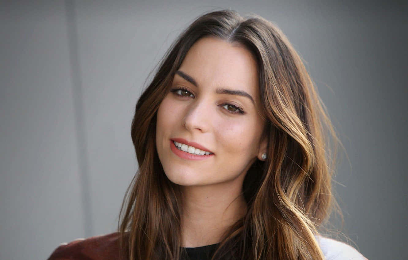 Genesis Rodriguez 桌布 壁紙 — 一位微笑女士的close-up肖像，她擁有長長嘅棕色頭髮同珍珠耳環，背景係中性灰色。