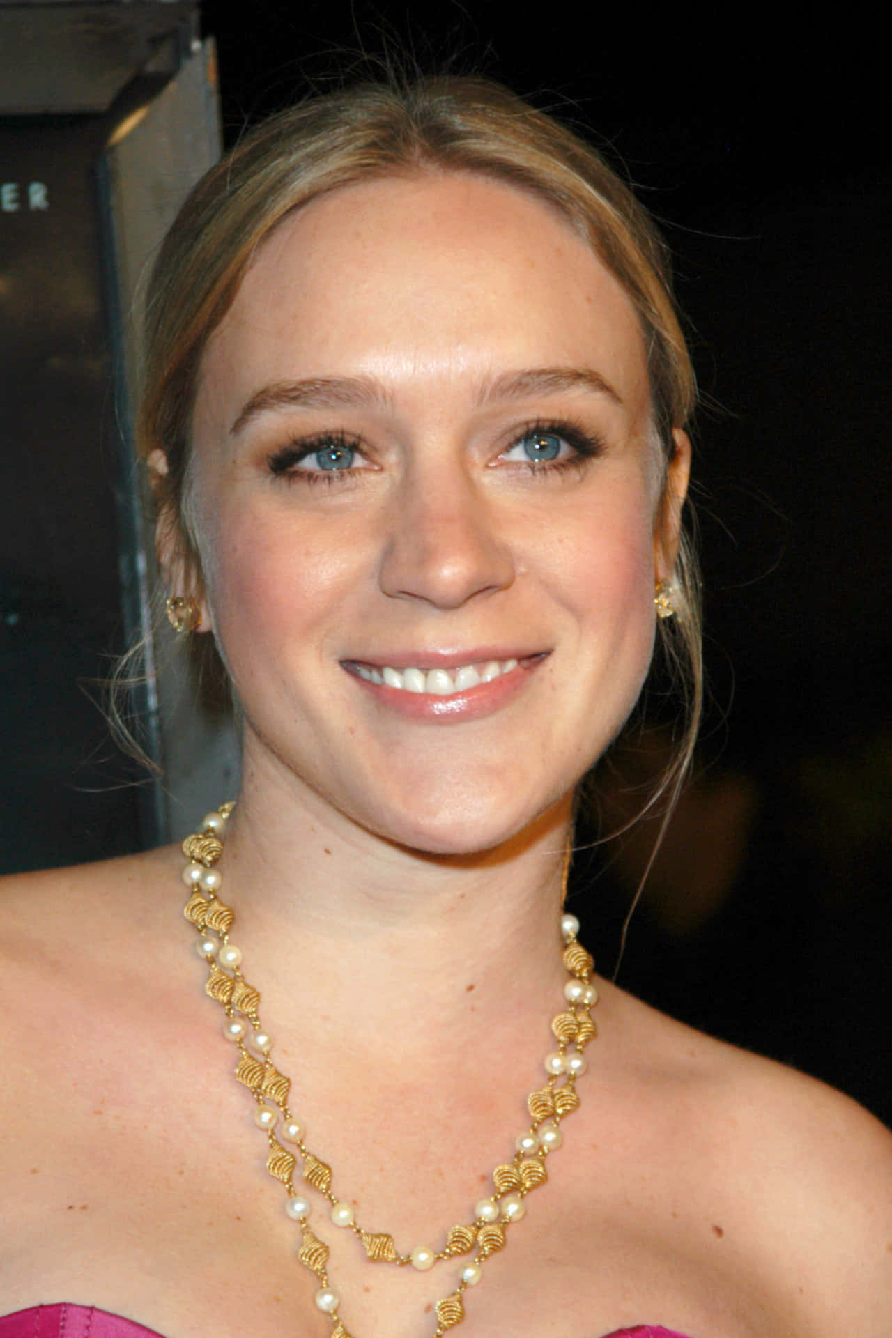 Chloë Sevigny 桌布 壁紙 — 一位微笑女士的近距離肖像，藍色眼睛，戴著金和珍珠項鍊，穿著粉紅色連衣裙。