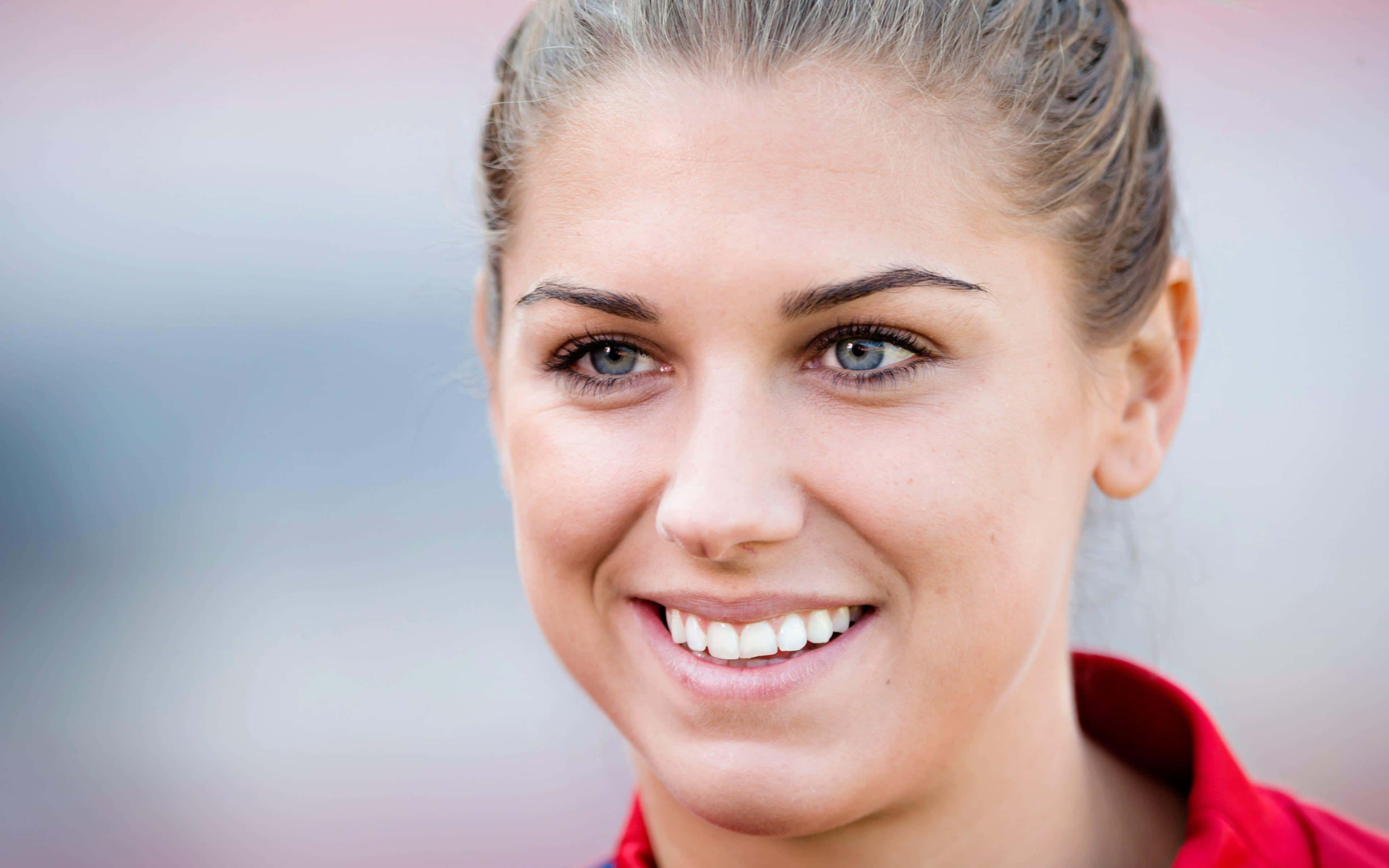 Alex Morgan 桌布 壁紙 — 一位微笑女性嘅特寫肖像，佢有藍色眼睛同金髮束起，著住紅色恤衫。