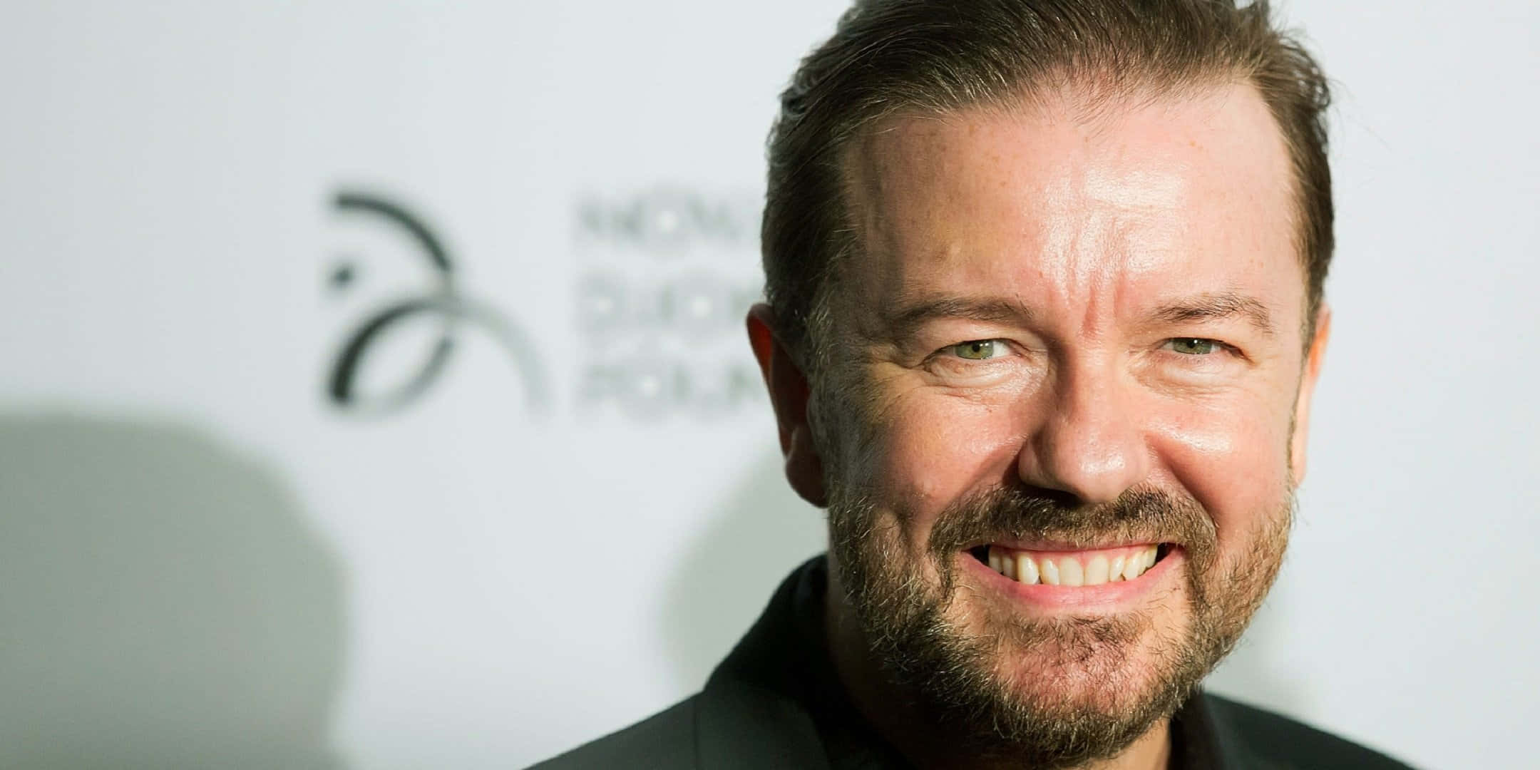 ricky gervais 桌布 壁紙 — 一位微笑留鬍鬚男士穿著黑色西裝，在白色背景前帶有標誌的特寫照片。