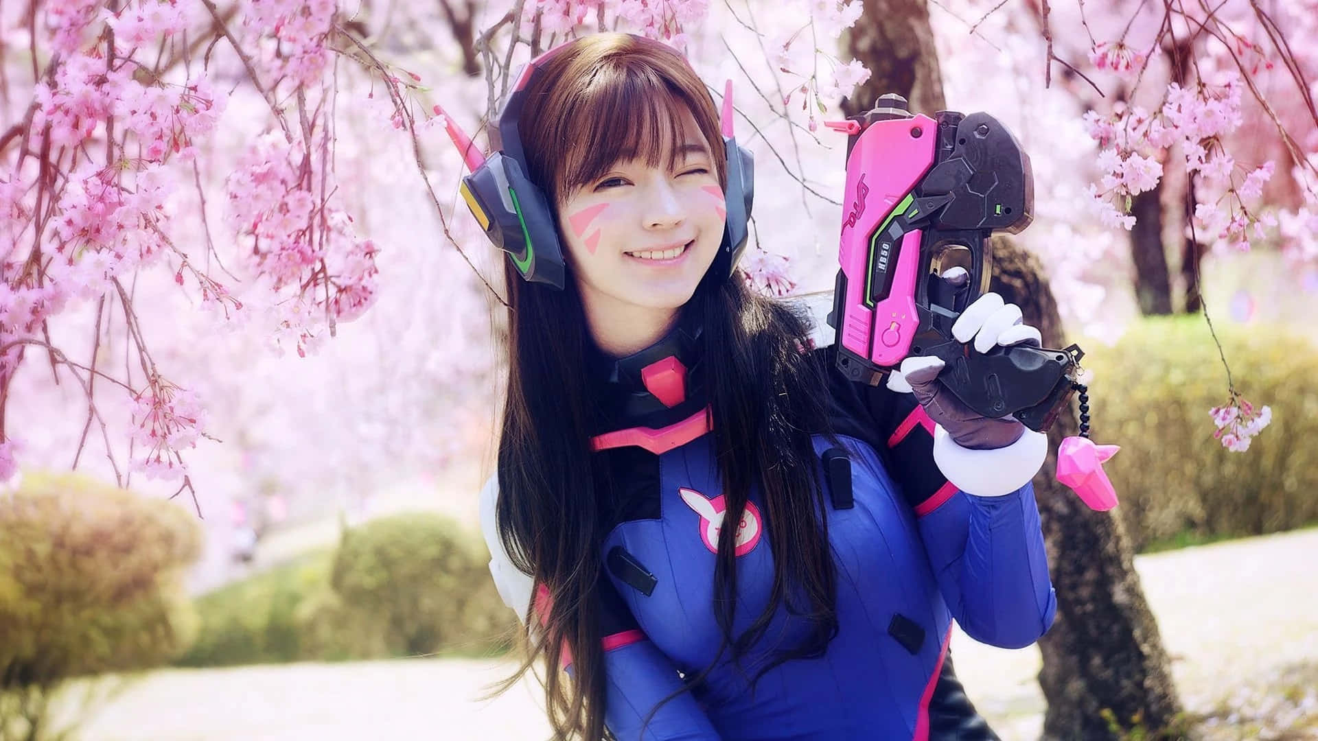 Cosplay Anime Kawaii 桌布 壁紙 — 一位微笑的Cosplayer穿著藍色和粉紅色的Overwatch服裝，在盛開的櫻花下拿著武器擺姿勢。