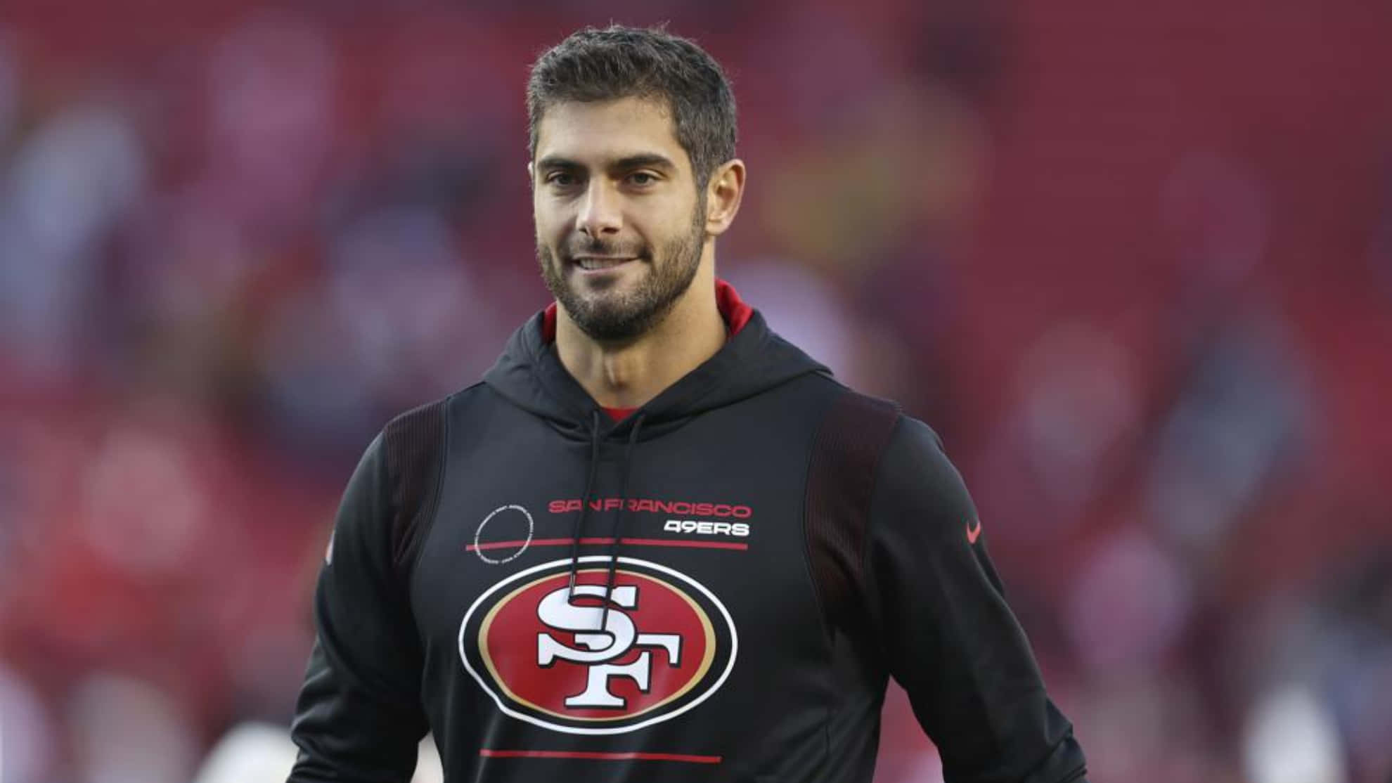 Jimmy Garoppolo 桌布 壁紙 — 一位微笑的三藩市49人球員穿著黑色球隊連帽衫，自信地站在模糊的球場上。