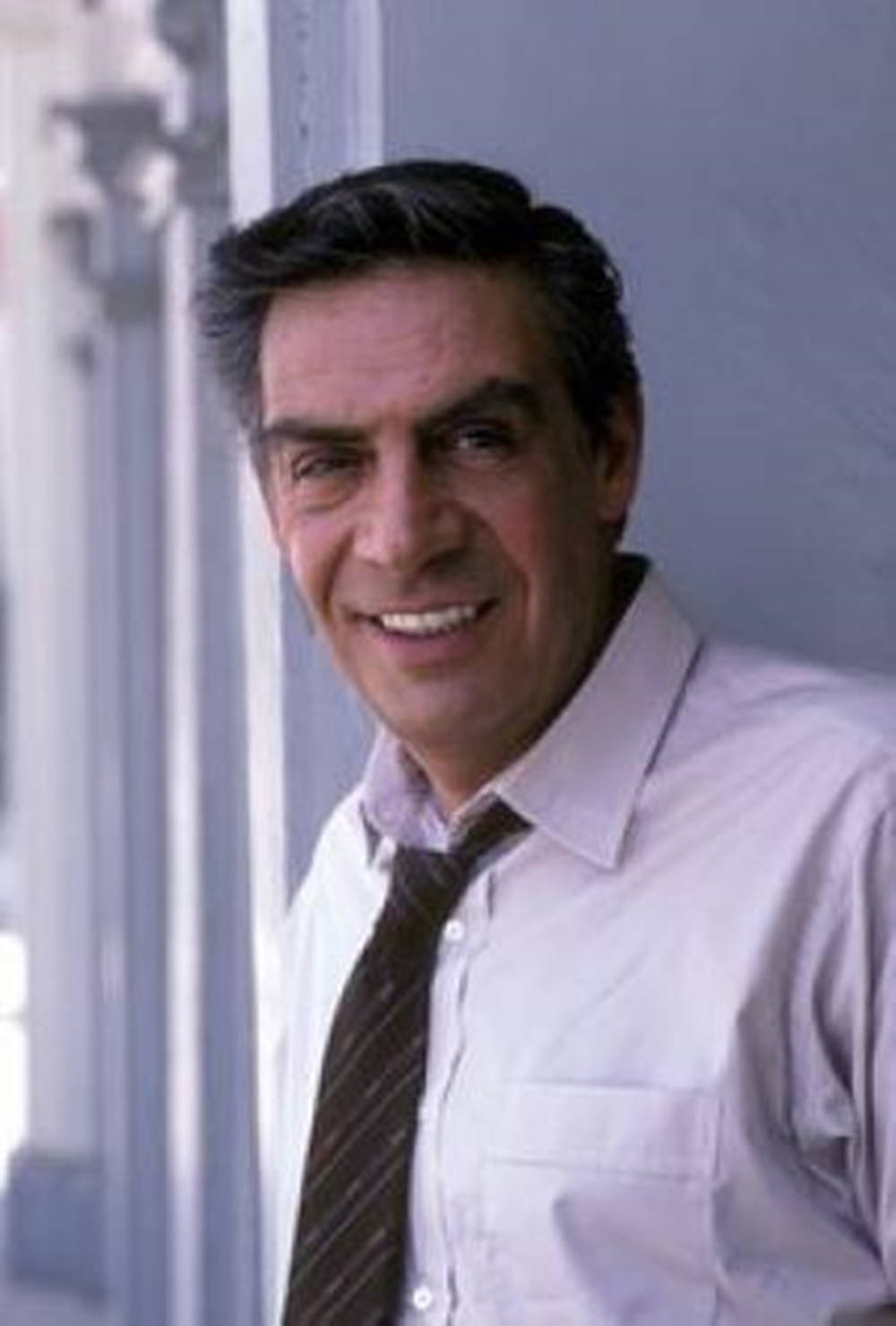 Actor Jerry Orbach Drake Houseman Dirty Dancing 壁紙 — 一位微笑的中年男子穿著淺色襯衫和條紋領帶，在柔和日光中靠著白色柱子。