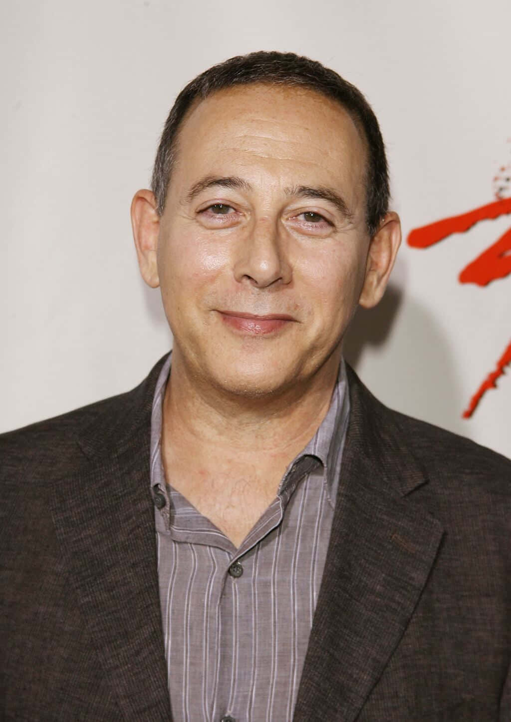 Paul Reubens 桌布 壁紙 — 一位微笑的中年男子身穿深色西裝外套和條紋襯衫，在白色背景上帶有紅色筆觸的特寫肖像。