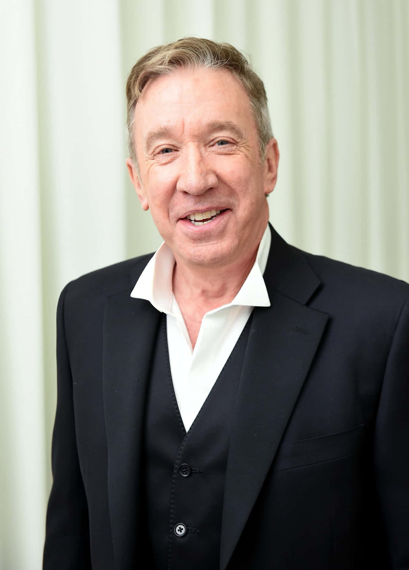 Tim Allen 桌布 壁紙 — 一位微笑的中年男子身穿黑色西裝和白色襯衫，站在柔和的奶油色窗簾背景前。