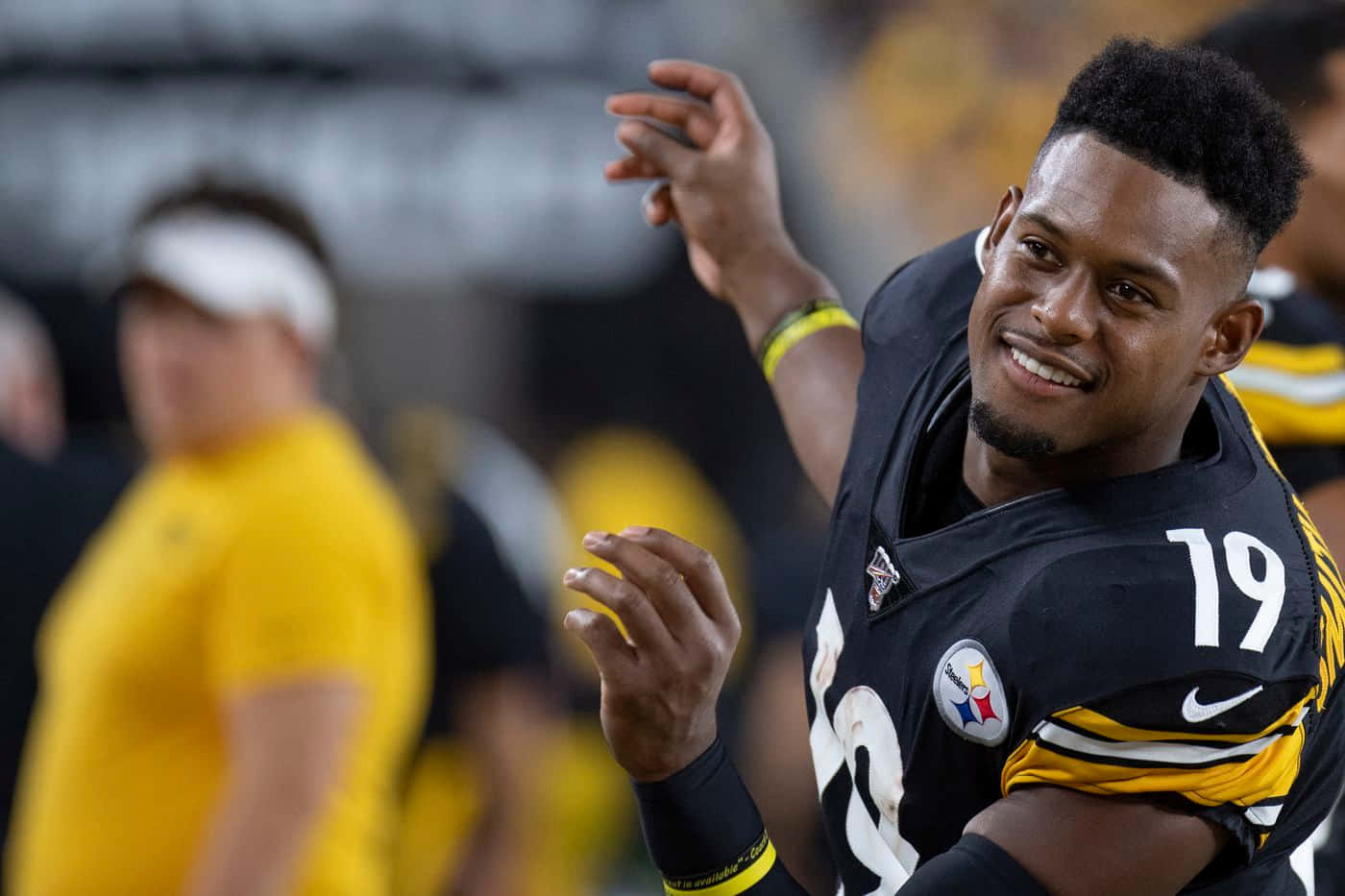Juju Smith Schuster 桌布 壁紙 — 一位微笑的匹茲堡鋼人隊美式足球員身穿19號球衣，在日間比賽中向球迷揮手。