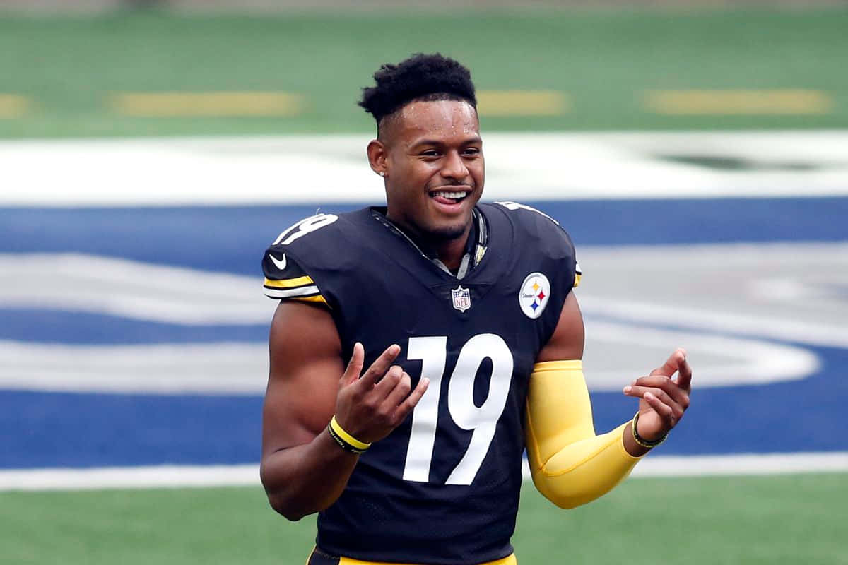 Juju Smith Schuster 桌布 壁紙 — 一位微笑的匹茲堡鋼人隊美式足球員，身穿19號球衣，在帶有藍白標記的綠色球場上慶祝。