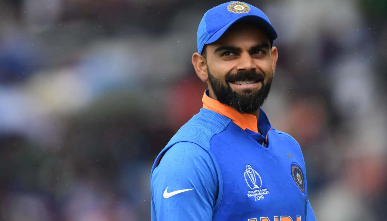 Virat Kohli 桌布 壁紙 — 一位微笑的印度板球隊隊長，身穿藍色隊服，配橙色領和帽子，自信地站在下雨的場地上。