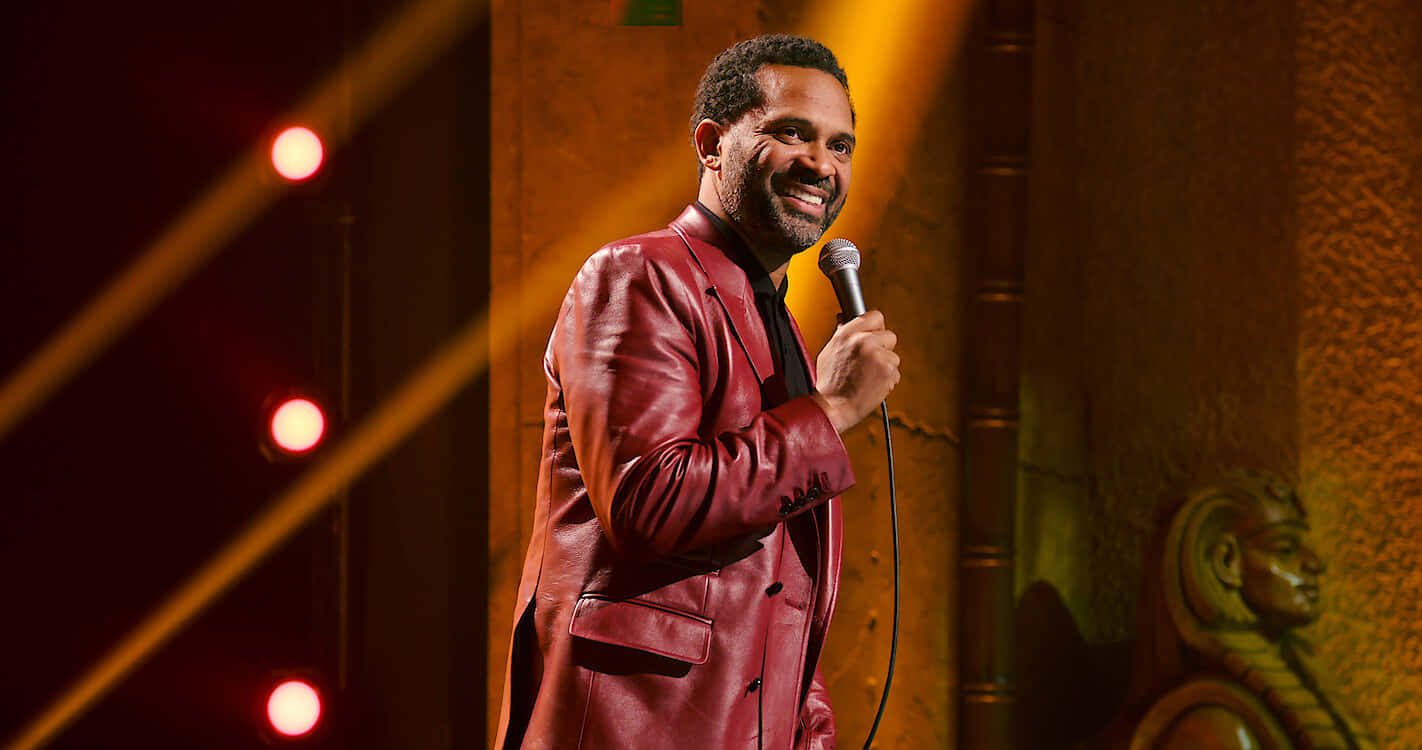 Mike Epps 桌布 壁紙 — 一位微笑的喜劇演員穿著紅色皮夾克，在溫暖的舞台燈光下握著麥克風，背景有發光的聚光燈。