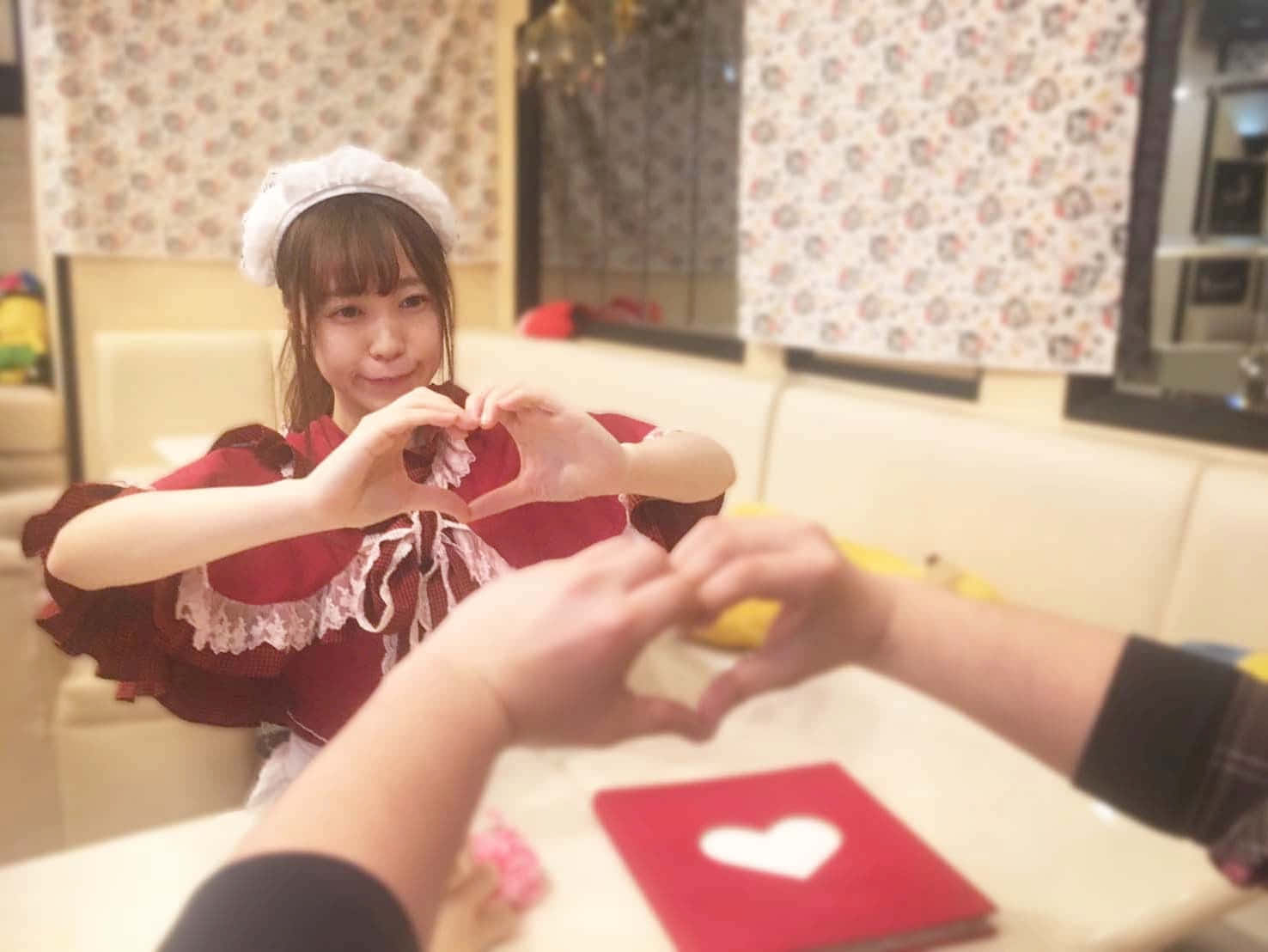 Otaku Culture 桌布 壁紙 — 一位微笑的女傭穿著紅色連衣裙，隔著桌子與顧客用手比出心形，顧客也做出同樣的手勢。
