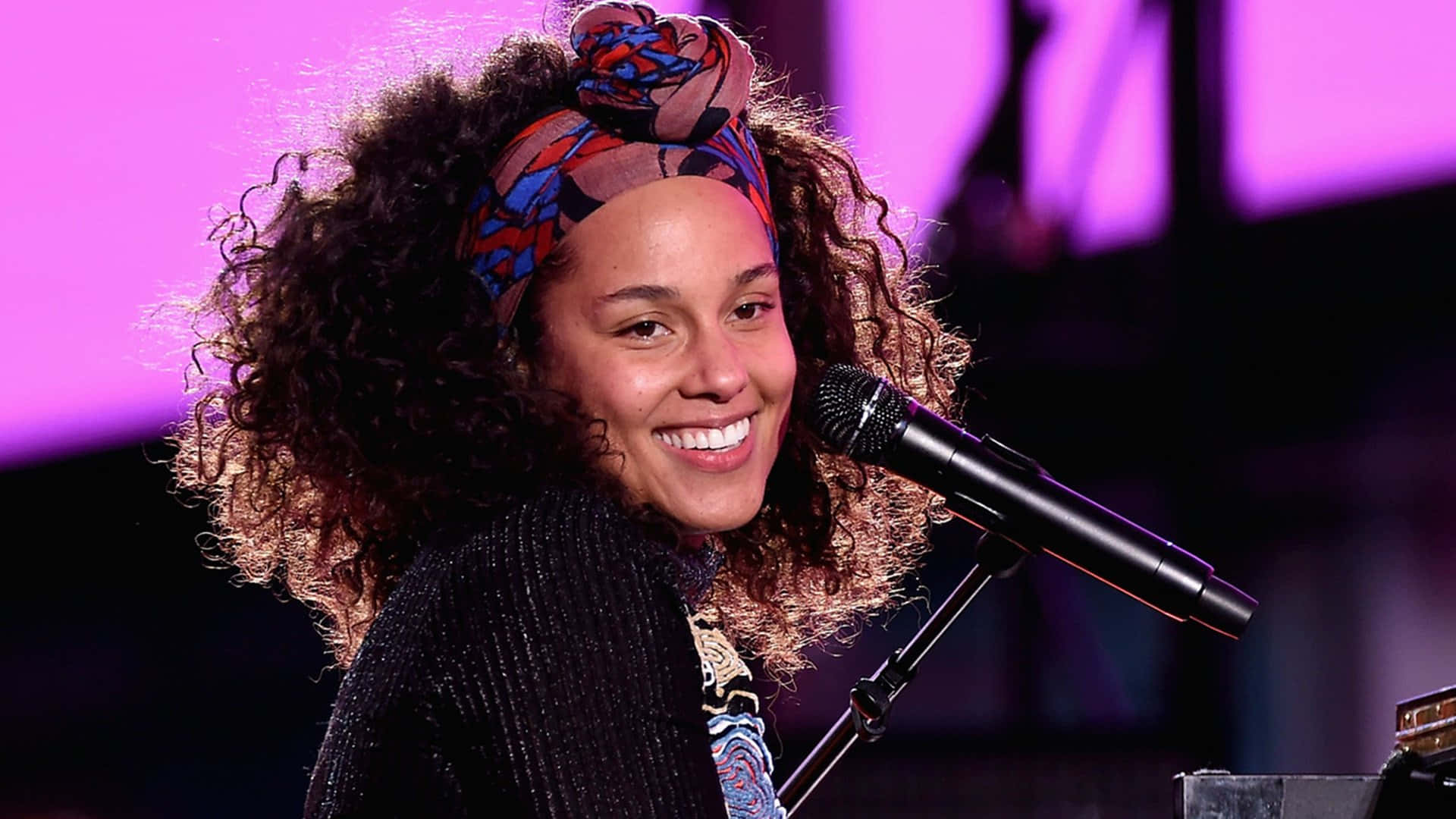 Alicia Keys 桌布 壁紙 — 一位微笑的女士，捲髮，戴著圖案頭巾，在紫色舞台燈光下對著麥克風唱歌。