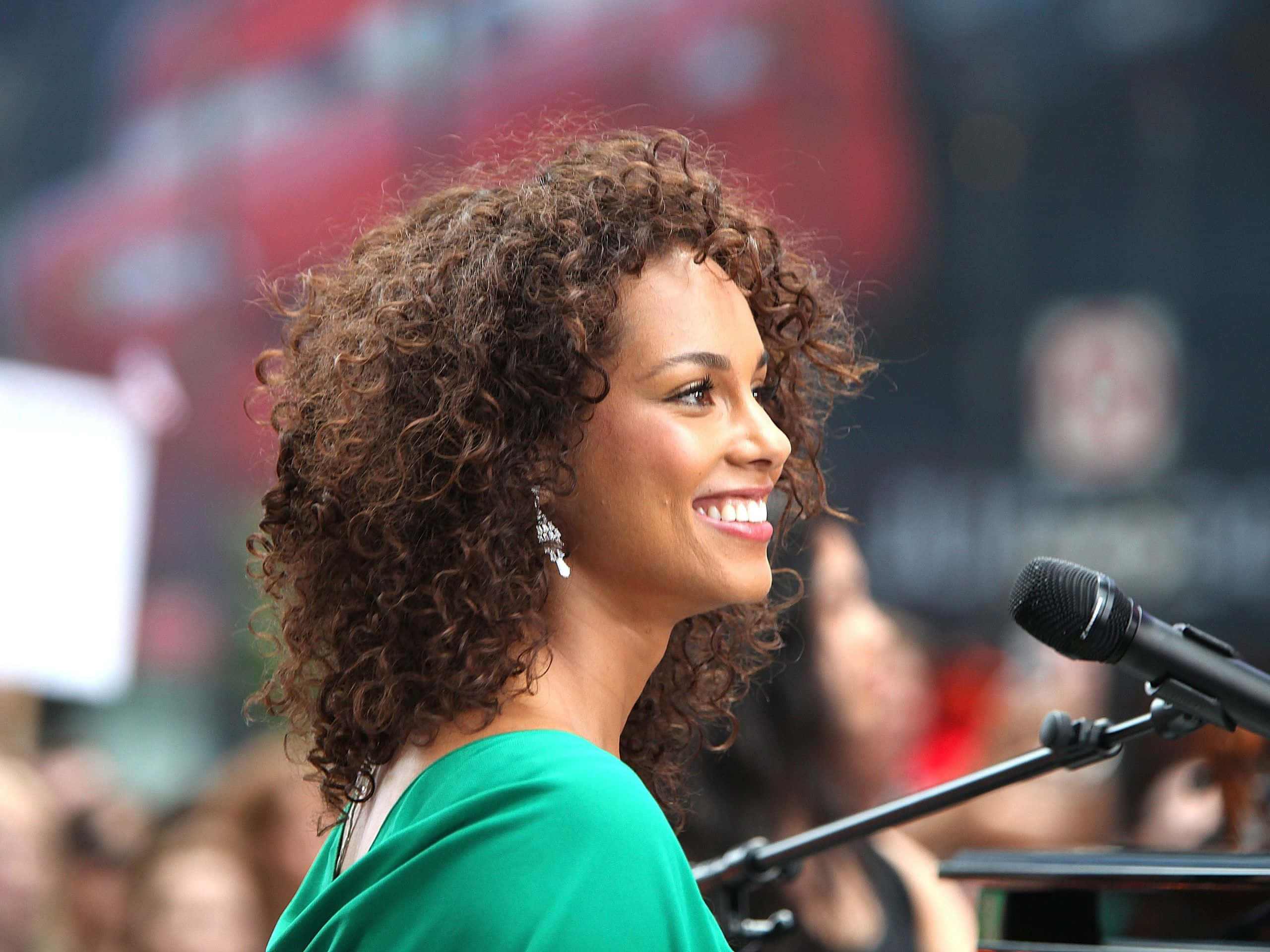Alicia Keys 桌布 壁紙 — 一位微笑的女士，棕色捲髮，穿著綠色連衣裙，站在舞台上的麥克風前，身後是模糊的人群。