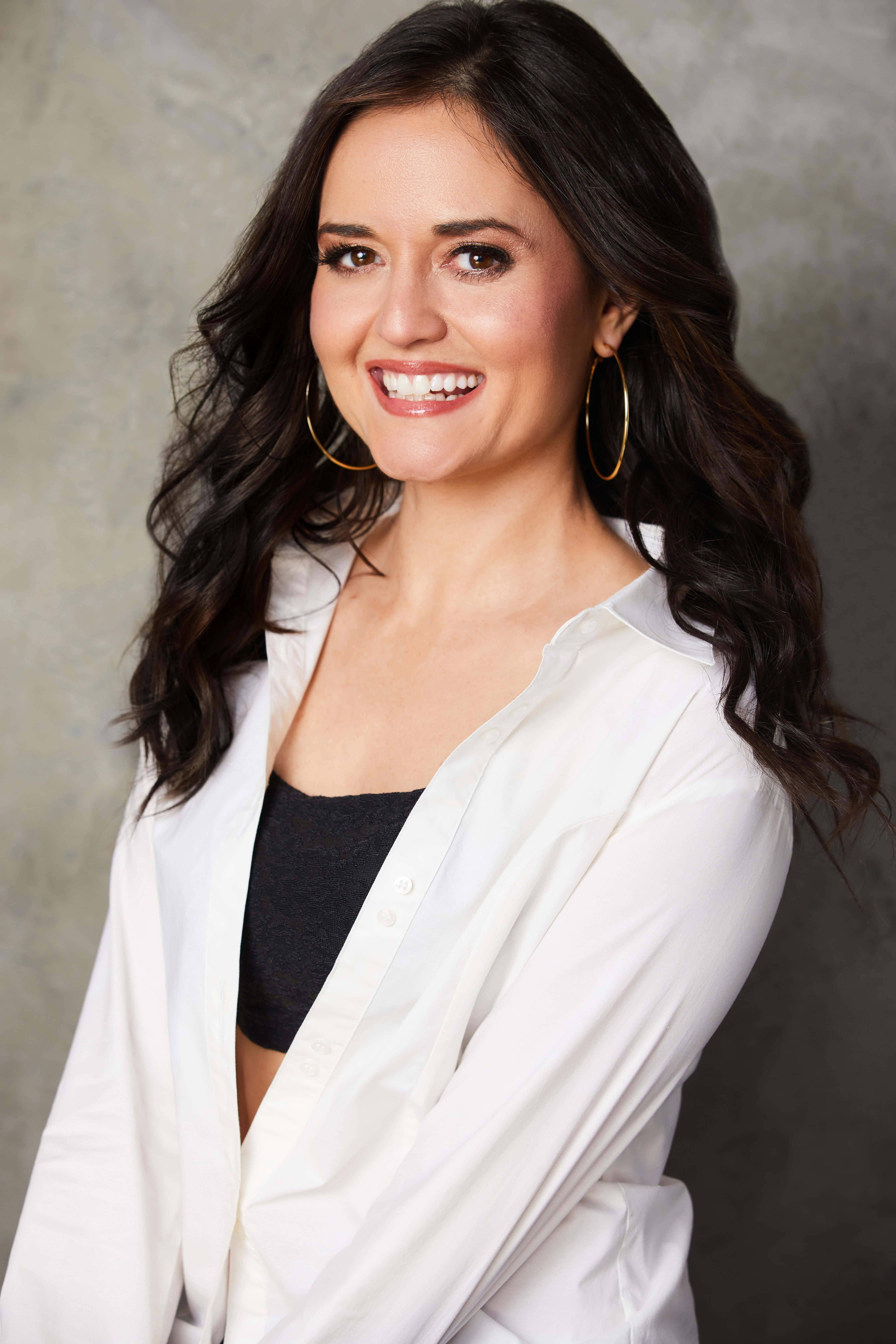 Danica Mckellar 桌布 壁紙 — 一位微笑的女士，深色波浪頭髮，戴著金色圓環耳環，在紋理灰色背景前，黑色上衣外穿白色襯衫。
