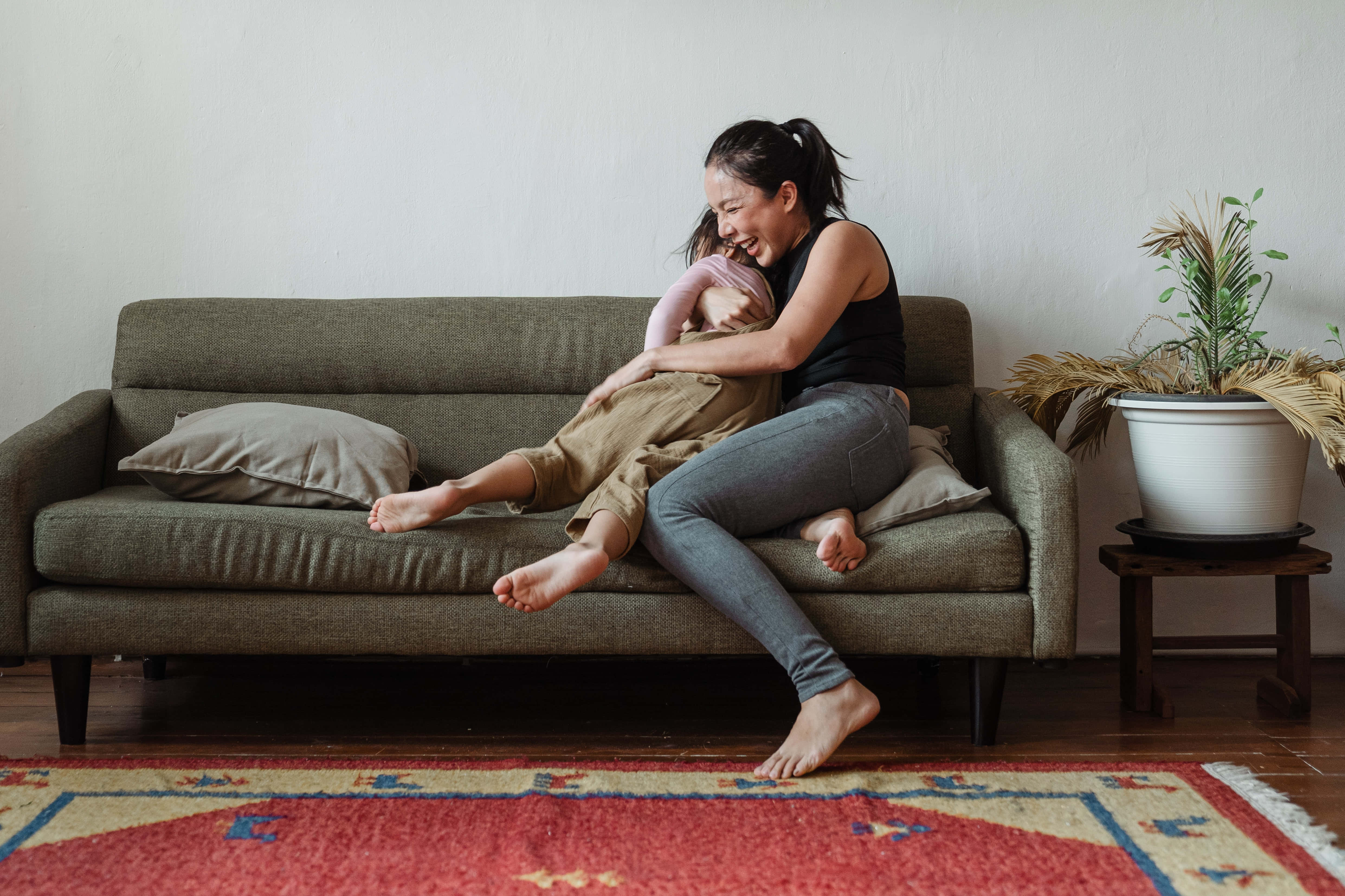 Mom 桌布 壁紙 — 一位微笑的女士在舒適的綠色沙發上抱著一個熟睡的嬰兒，旁邊有一盆植物和一塊圖案地毯。