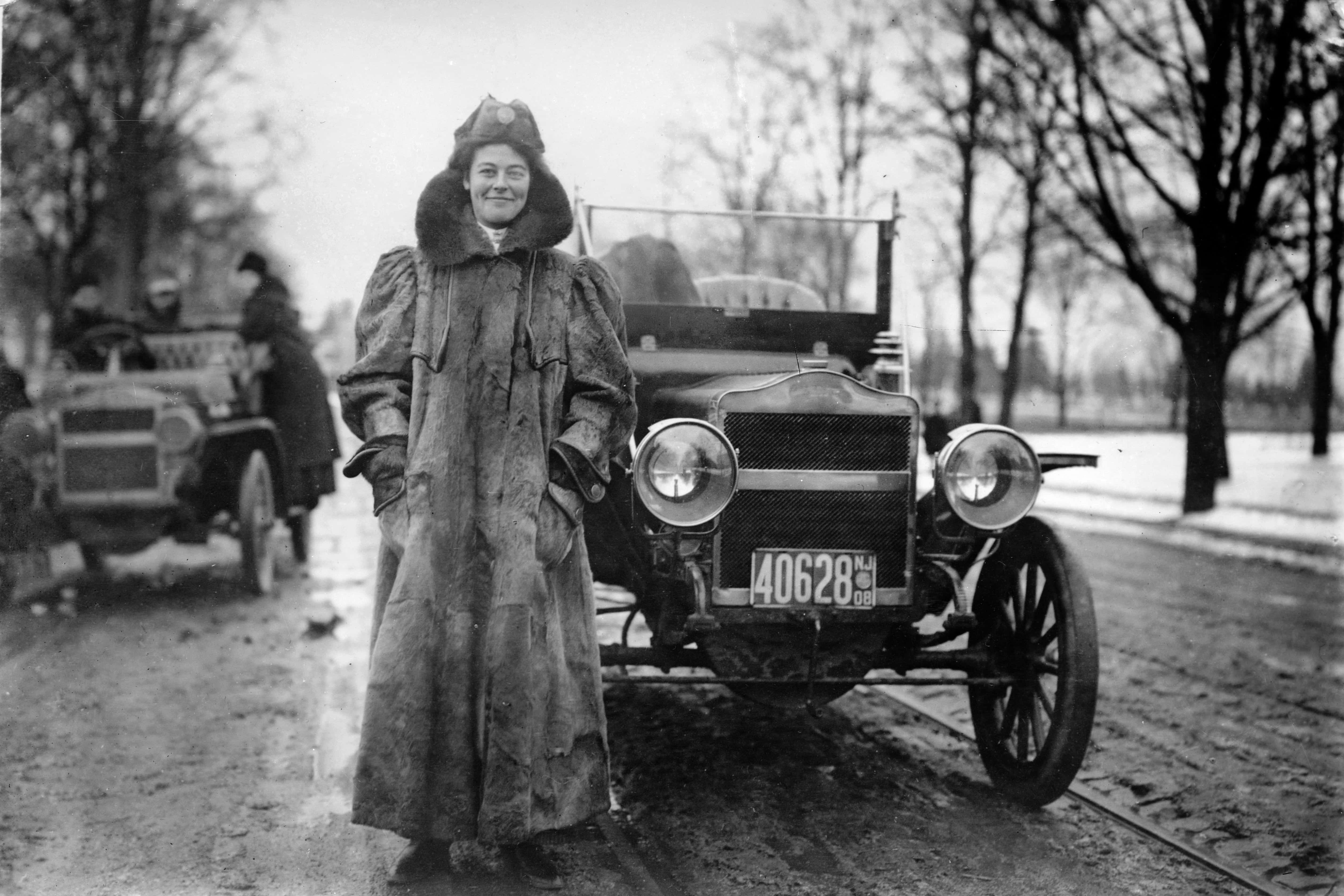Black And White Vintage Car 桌布 壁紙 — 一位微笑的女士穿著毛邊大衣，自豪地站在一輛復古福特Model T旁邊，背景是雪地路邊和光禿禿的樹木。