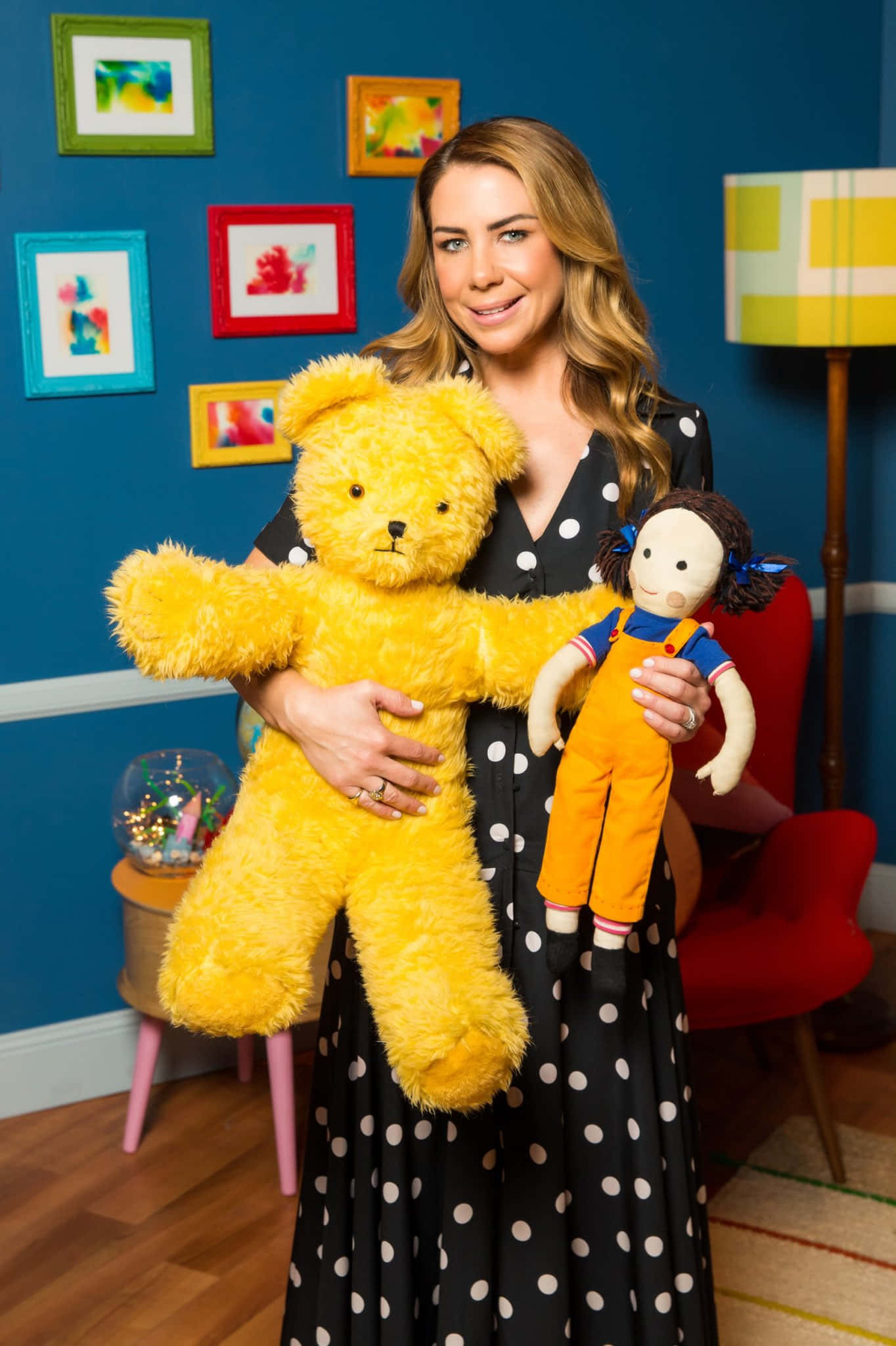 Kate Ritchie 桌布 壁紙 — 一位微笑的女士穿著波點裙，在色彩繽紛的裱框藝術牆前抱著一隻大型黃色泰迪熊和一個小娃娃。