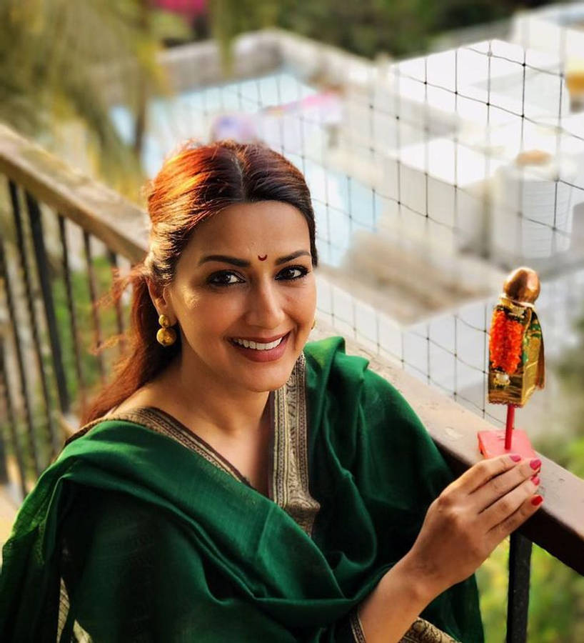 Gudi Padwa 桌布 壁紙 — 一位微笑的女士穿著綠色紗麗，在陽台上手持裝飾過的迪亞，背景是模糊的花園和泳池。