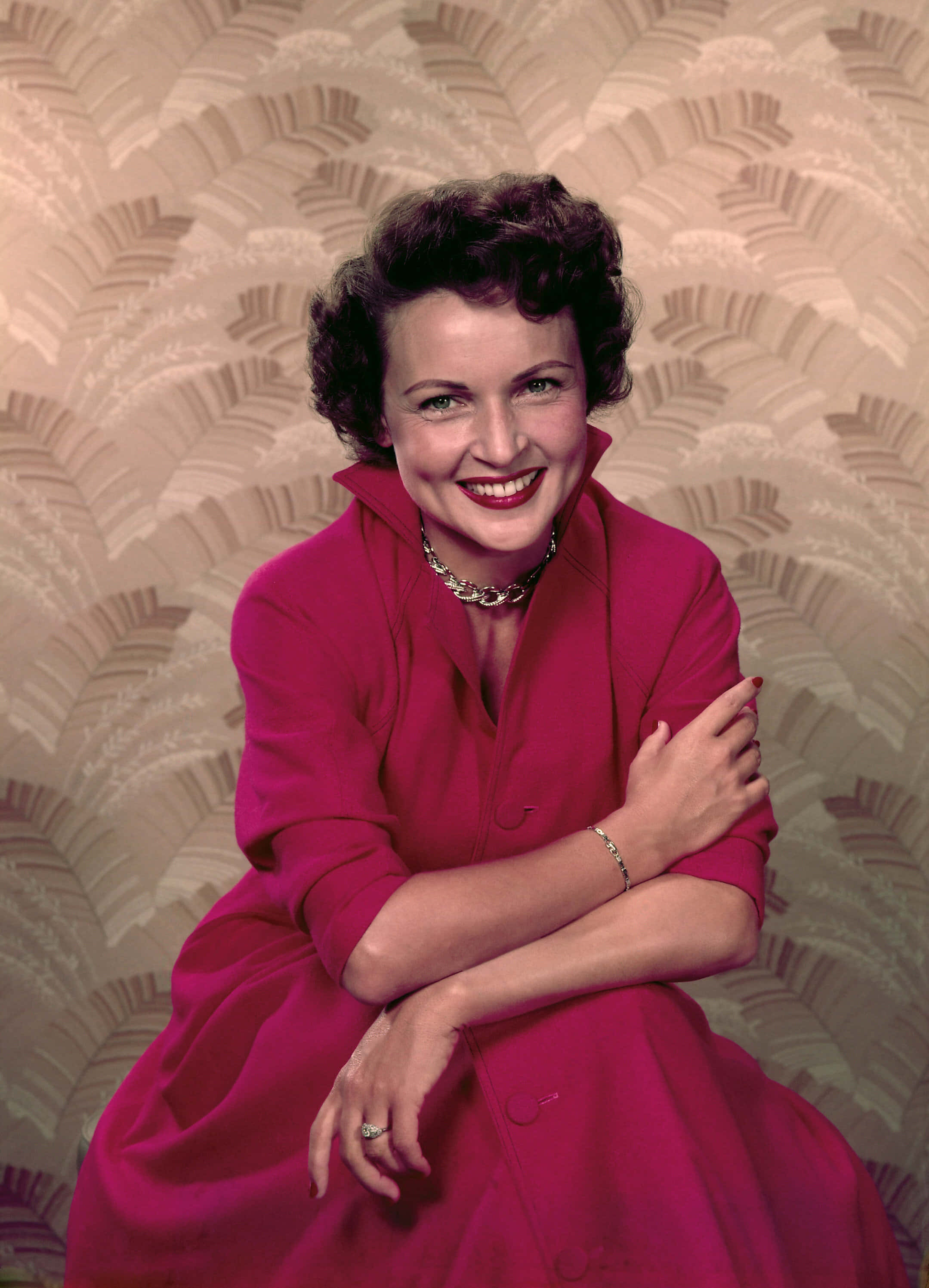 Betty White 桌布 壁紙 — 一位微笑的女士穿著鮮豔的粉紅色連衣裙，自信地擺姿勢，背景是帶有扇形圖案的紋理米色牆紙。