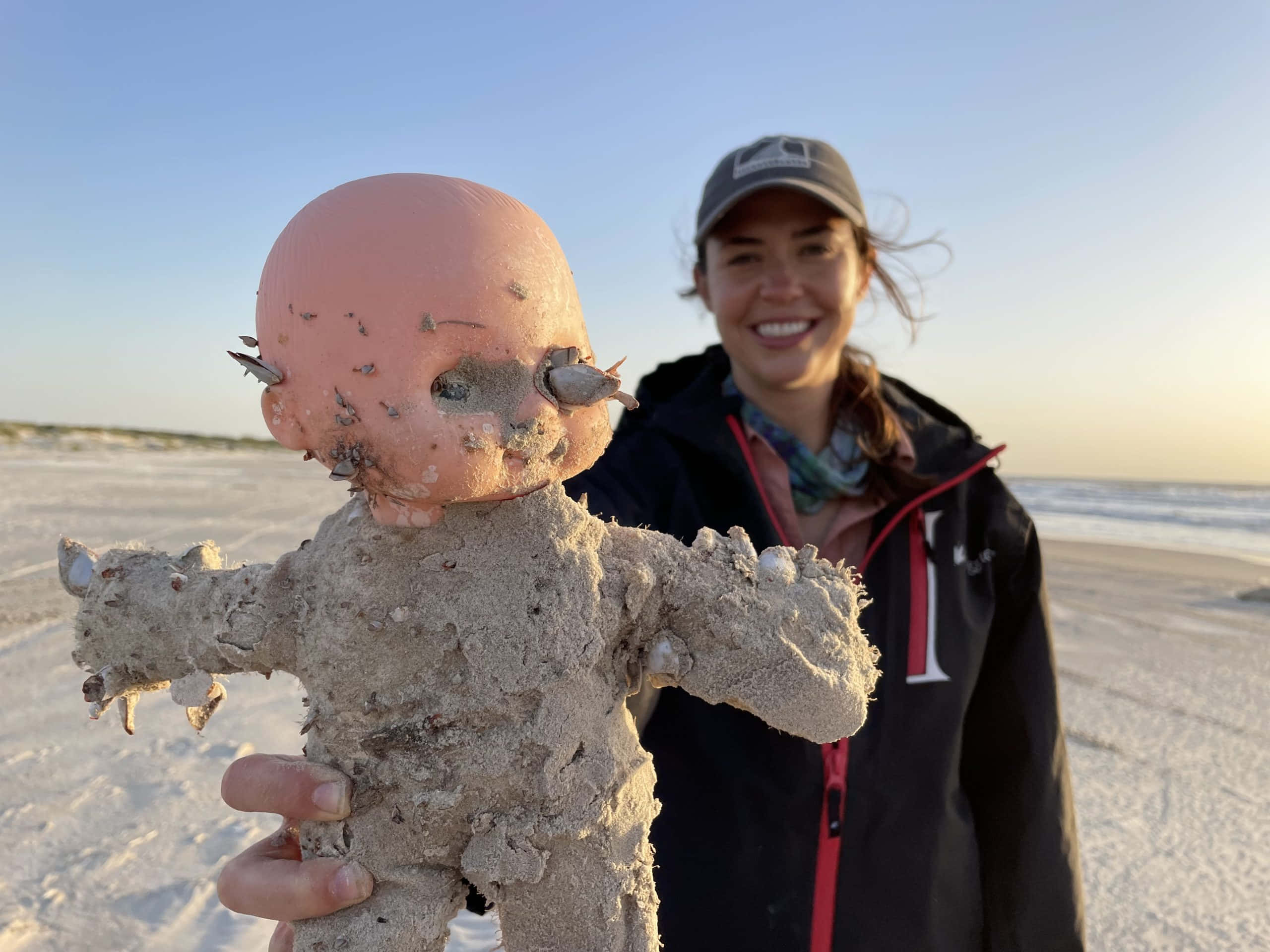 Dolls 桌布 壁紙 — 一位微笑的女子在海灘日落時分，抱著一個滿是沙粒、眼睛破損的洋娃娃。