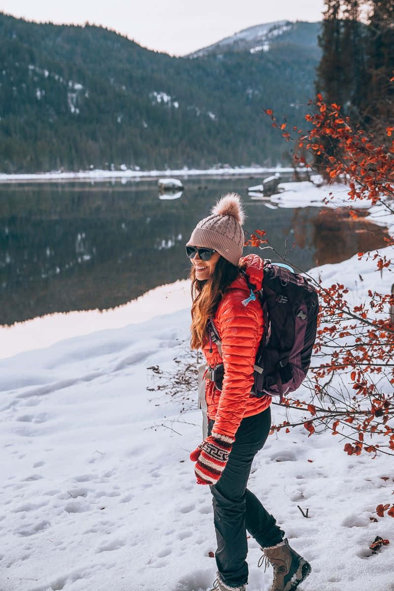 Winter Hiking 桌布 壁紙 — 一位微笑的女子穿著亮橙色外套和粉紅色無邊帽，在雪覆蓋的森林中行走，旁邊是平靜的湖泊，背景是白雪覆蓋的山脈。