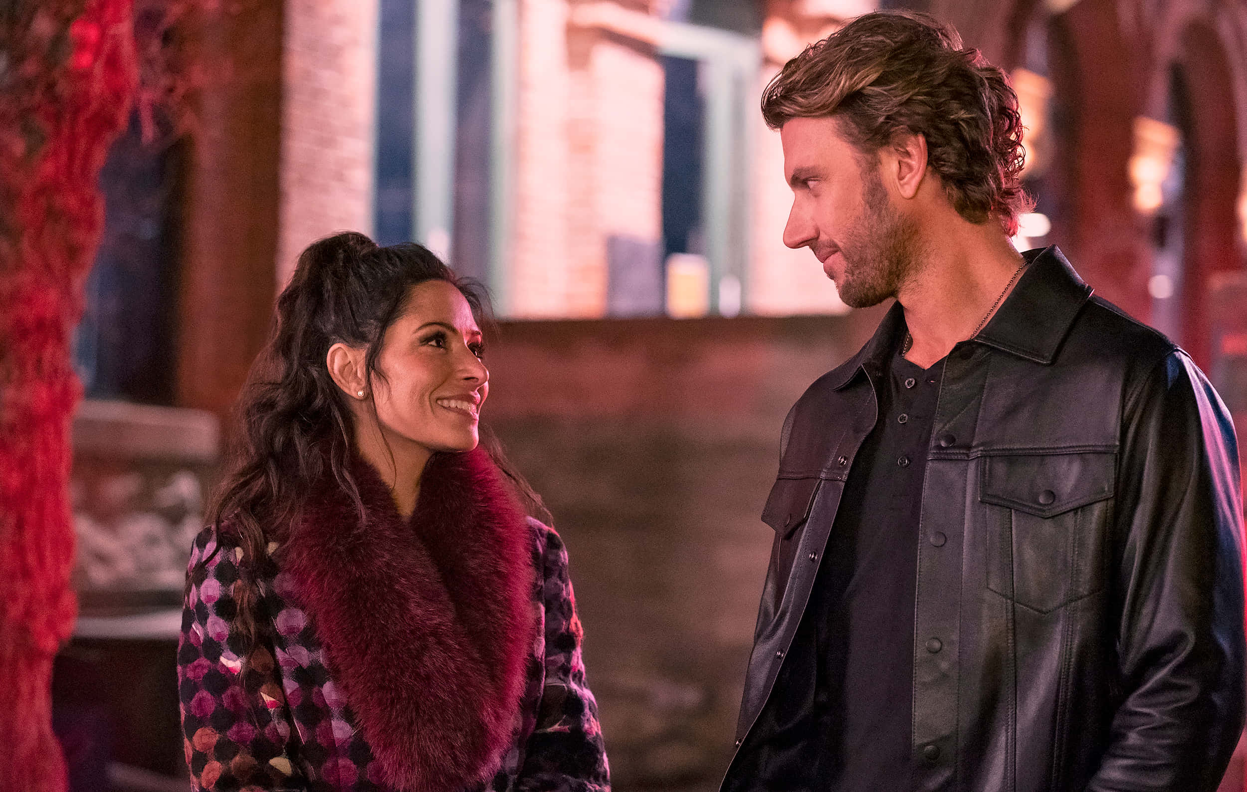 sarah shahi 桌布 壁紙 — 一位微笑的女子穿著毛領大衣，一位男子穿著皮褸，在溫暖的夜燈下分享親密時刻。
