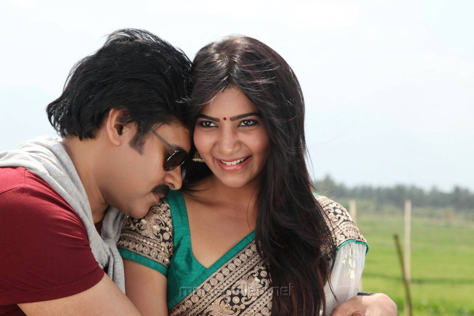 Pawan Kalyan 桌布 壁紙 — 一位微笑的女子穿著綠色和金色的紗麗，在明亮的綠色田野背景下擁抱一位穿著紅色襯衫和太陽眼鏡的男子。