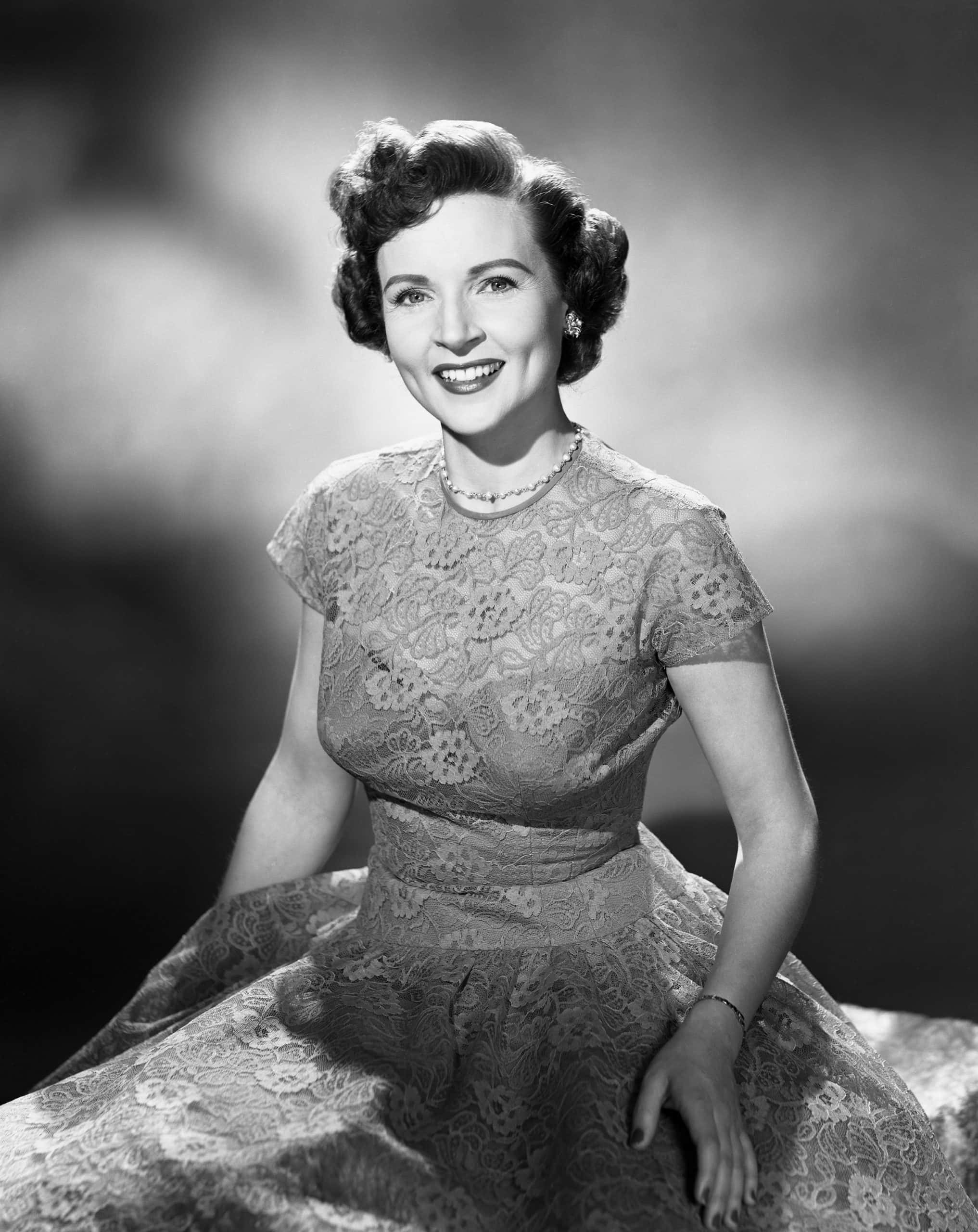 Betty White 桌布 壁紙 — 一位微笑的女子穿著蕾絲裙，在柔和模糊的攝影棚背景前優雅地擺姿勢。