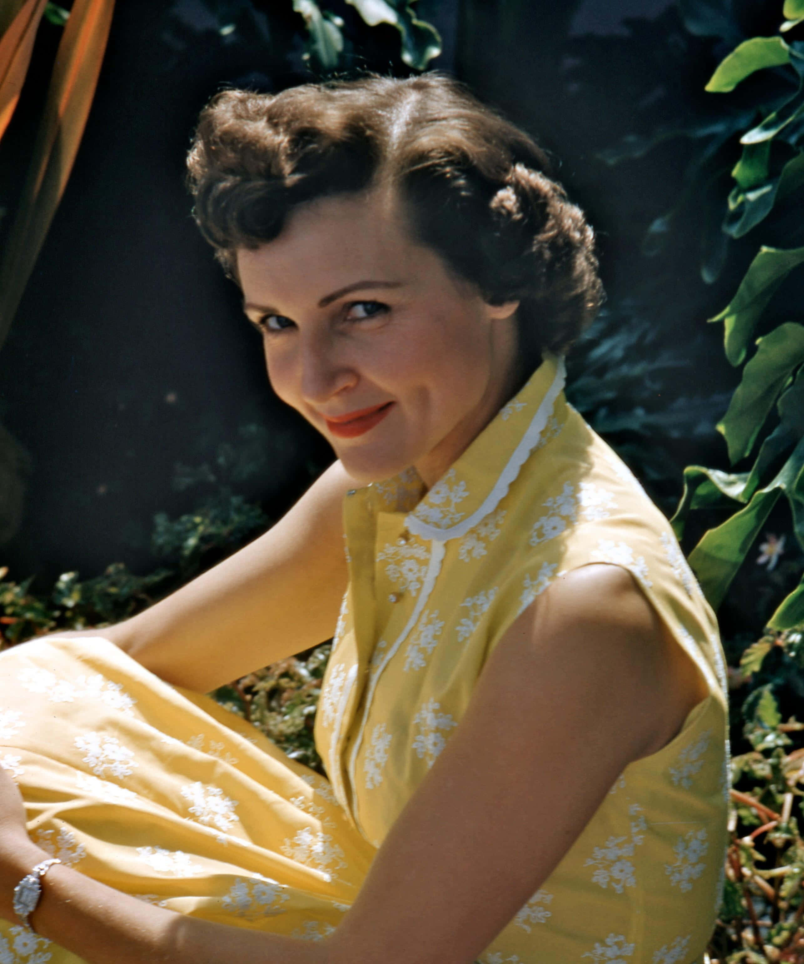 Betty White 桌布 壁紙 — 一位微笑的女子穿著黃色花卉連衣裙，在明亮的陽光下坐在戶外，周圍環繞著茂密的綠色樹葉。