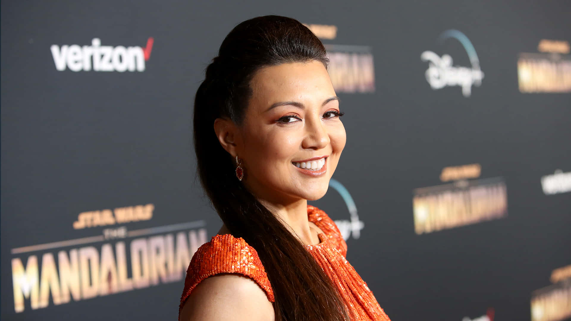 Ming Na Wen 桌布 壁紙 — 一位微笑的女子身穿橙色閃片連衣裙，在《星球大戰：曼達洛人》活動中，於品牌背景前擺姿勢。