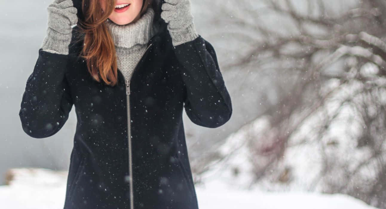 Winter Fashion 桌布 壁紙 — 一位微笑的女子身穿黑色外套和灰色手套，站在飄雪中，身後是積雪的樹木。