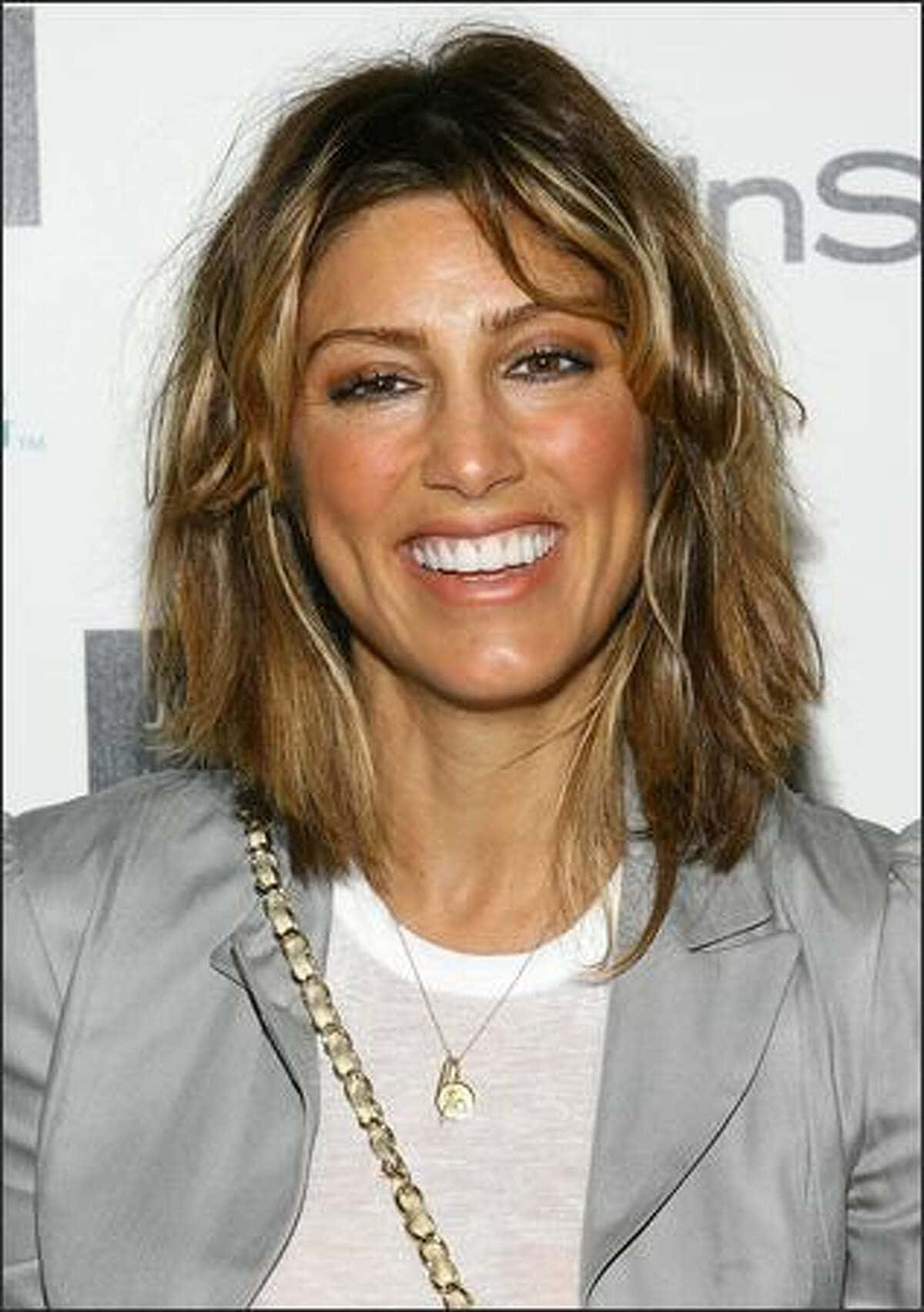 Jennifer Esposito 桌布 壁紙 — 一位微笑的女性，棕色亂髮，穿著灰色外套和金色鏈條項鍊，背景為白色。