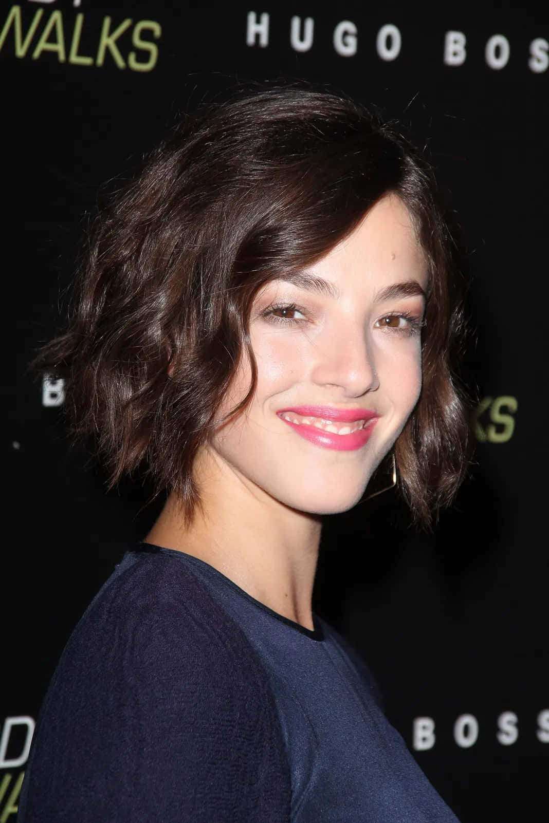 Olivia Thirlby 桌布 壁紙 — 一位微笑的女性，留著深色波浪短髮和亮粉紅色唇膏，在黑色背景帶有標誌的Hugo Boss活動中擺姿勢。