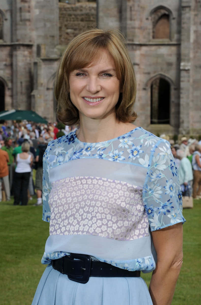 Fiona Bruce 桌布 壁紙 — 一位微笑的女性，身穿藍色花卉圖案上衣和淺藍色裙子，站在戶外一座歷史悠久的石造建築前，身後有一群人。