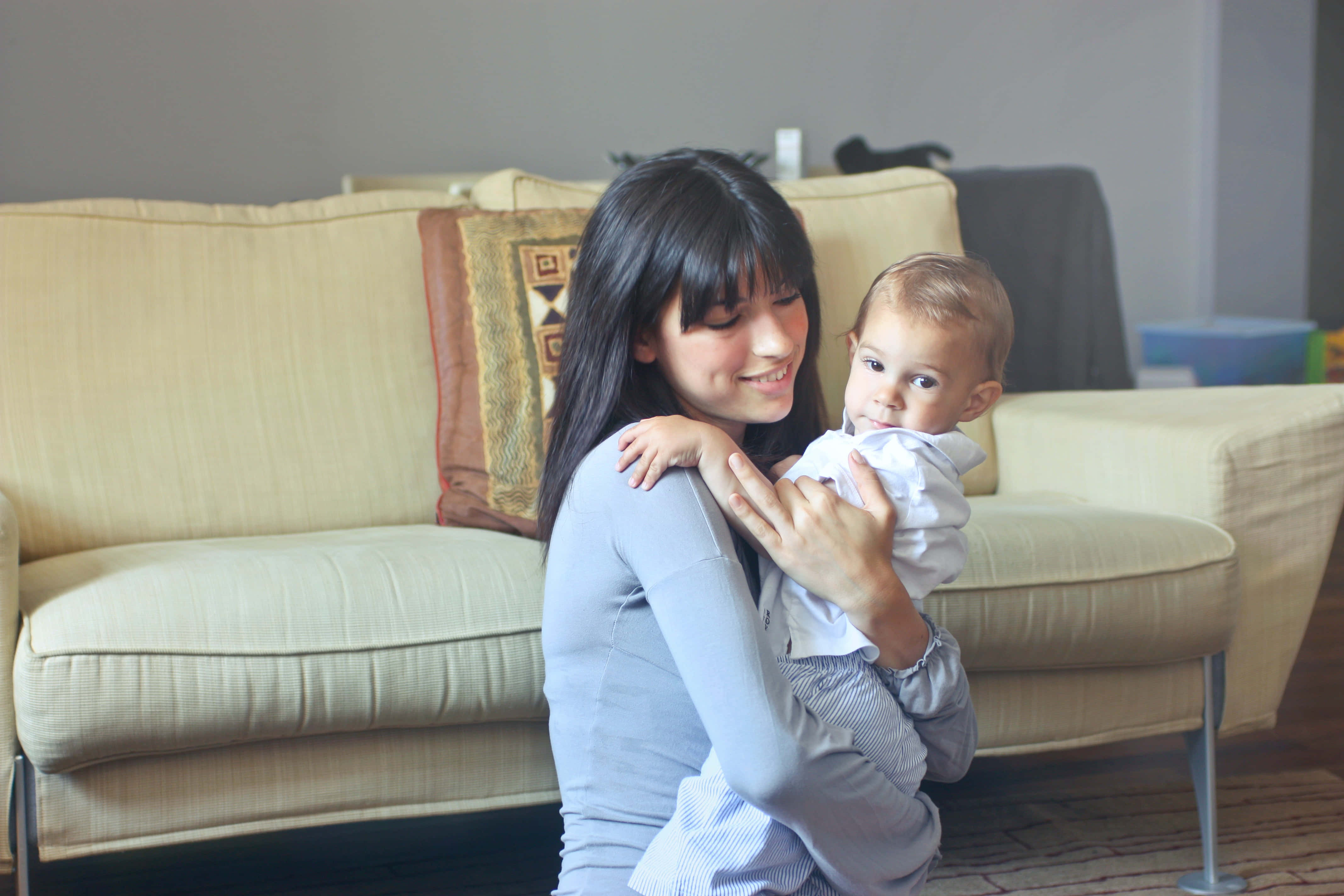 Mom 桌布 壁紙 — 一位微笑的女性在舒適的客廳中抱著嬰兒，客廳配有米色沙發和圖案抱枕。
