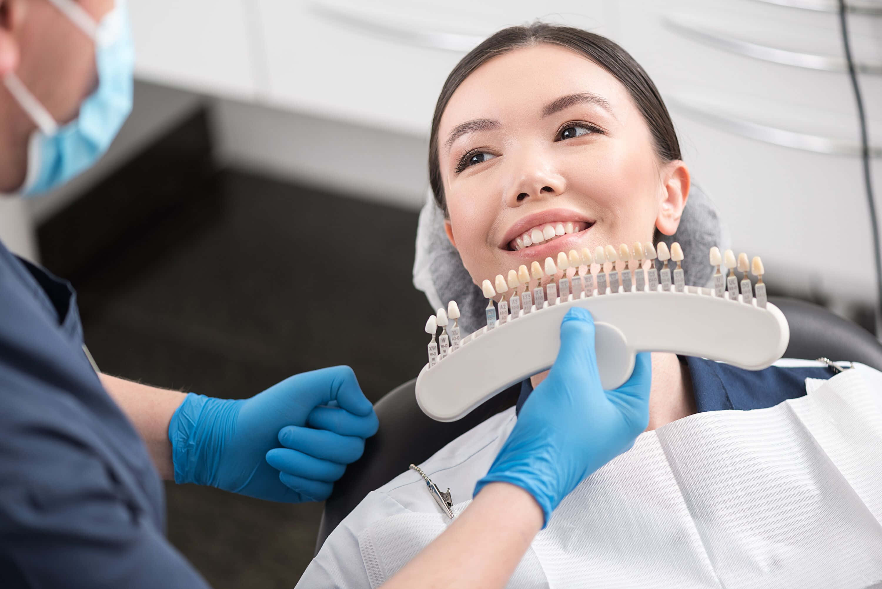 Teeth Whitening 桌布 壁紙 — 一位微笑的女性坐在牙科椅上，看著戴手套的牙醫在明亮的診所中拿著牙齒色調樣本。