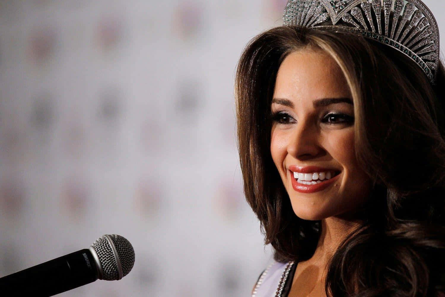 olivia culpo 桌布 壁紙 — 一位微笑的女性戴著閃亮的皇冠和禮服，對著模糊圖案背景前的麥克風說話。