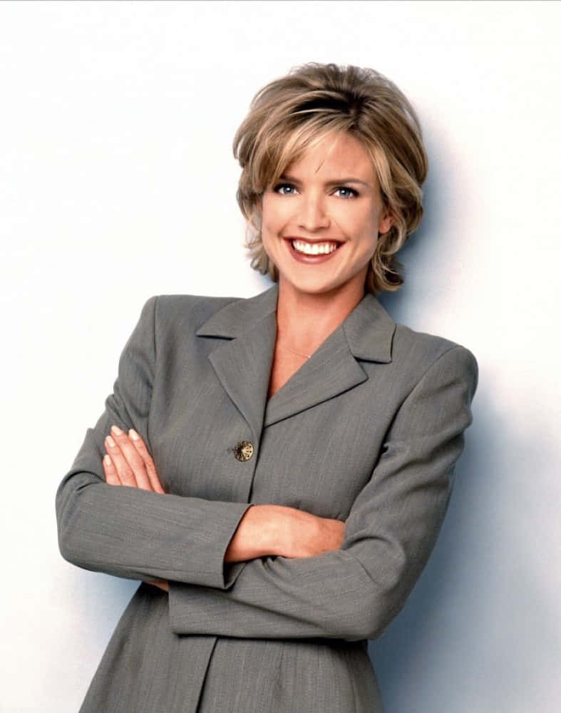 Courtney Thorne Smith 桌布 壁紙 — 一位微笑的女性穿著灰色西裝，自信地交叉雙臂站在純白色背景前。