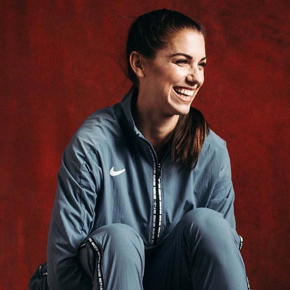 Alex Morgan 桌布 壁紙 — 一位微笑的女性穿著藍色Nike運動服，蹲在質感紅牆前，散發著歡樂的能量。