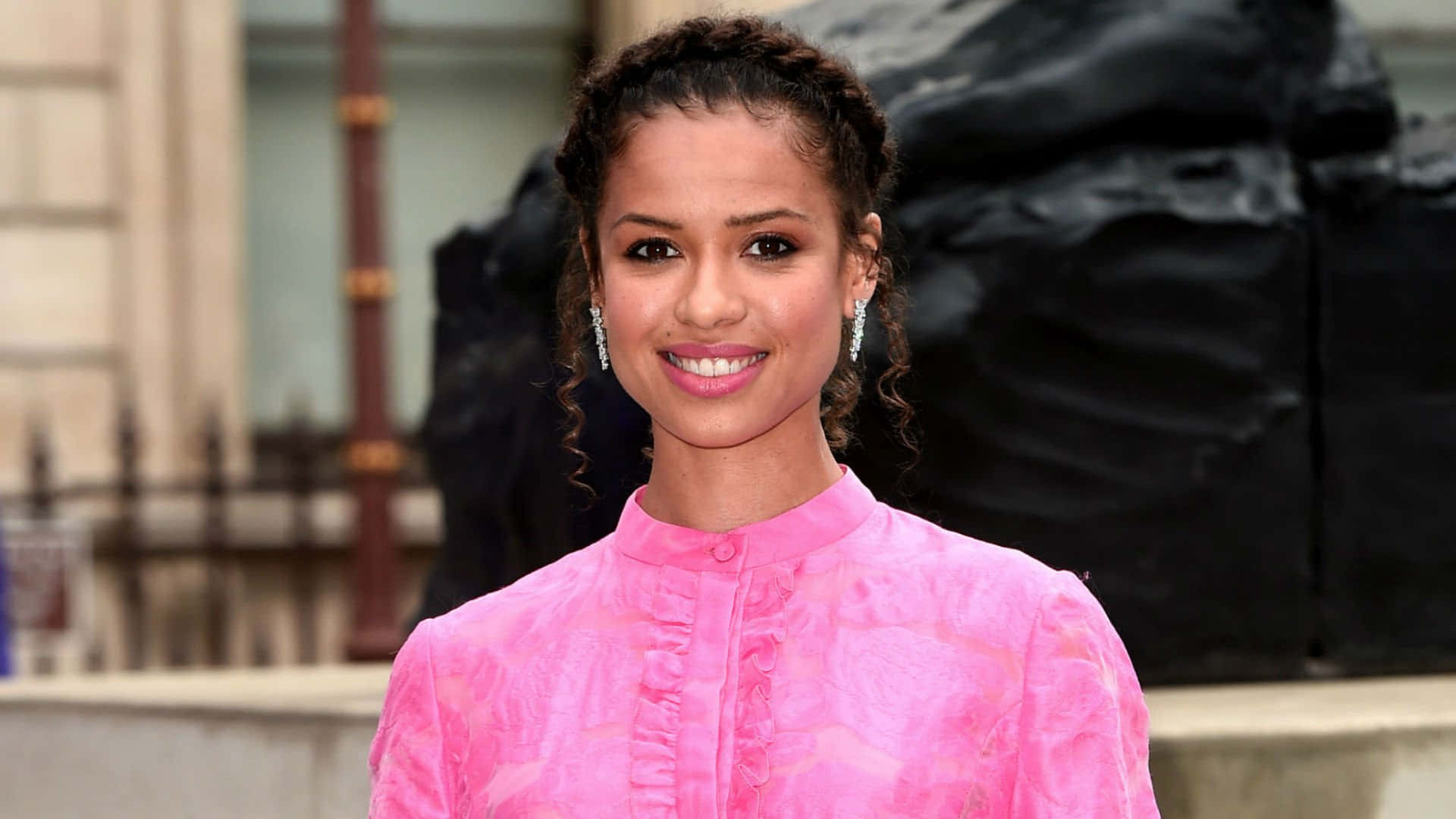 Gugu Mbatha-raw 桌布 壁紙 — 一位微笑的女性穿著鮮豔的粉紅色質感襯衫，站在深色雕塑和石造建築外牆前。