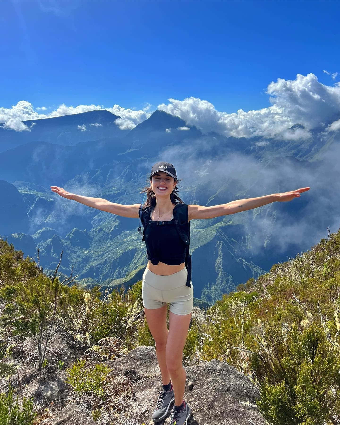 Hiking 桌布 壁紙 — 一位微笑的女性站在山脊上，雙臂張開，背景是霧氣繚繞的山峰和充滿活力的藍天。
