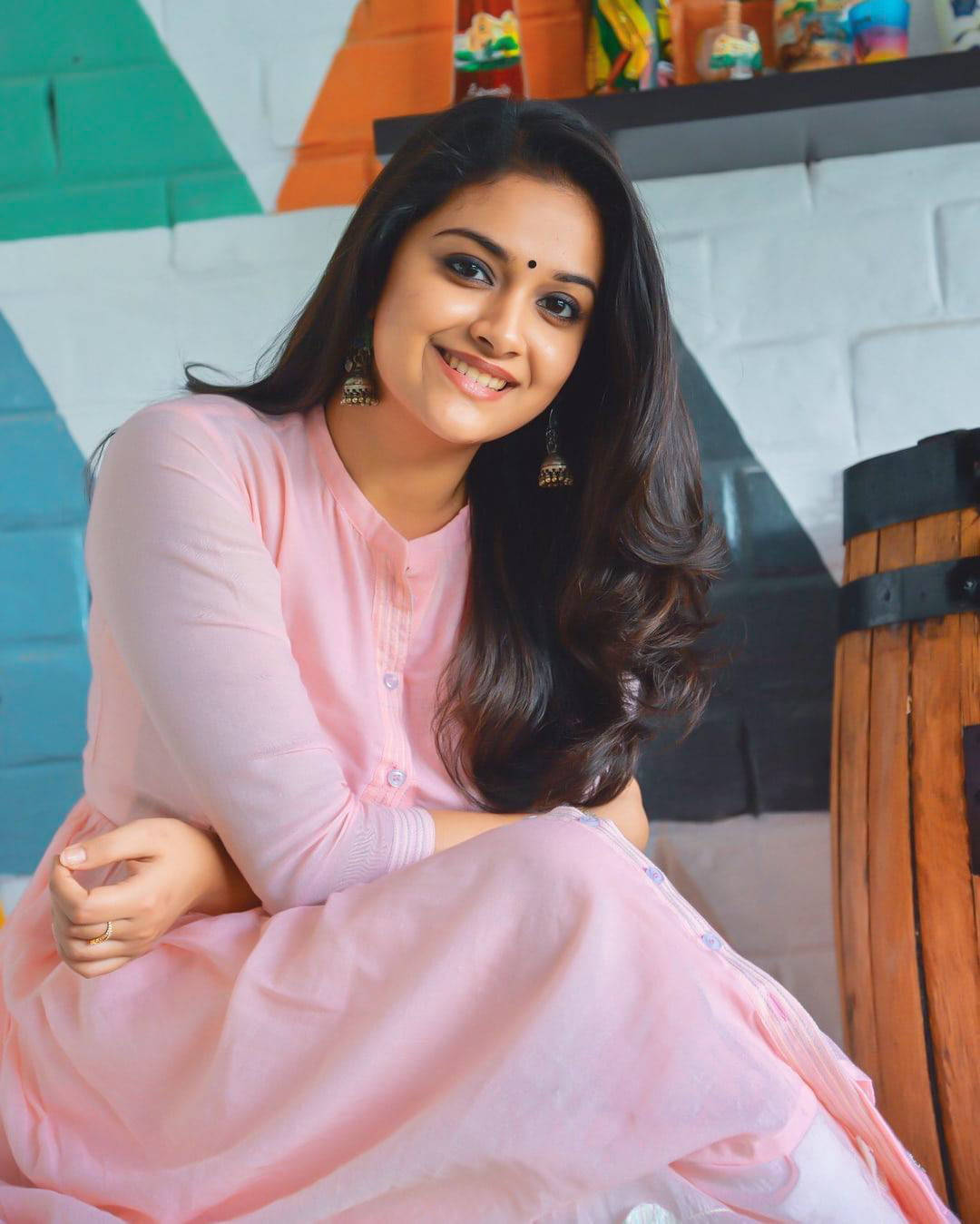 Tamil Actors HD 桌布 壁紙 — 一位微笑的女性身穿柔和粉紅色傳統服裝，優雅地坐在色彩繽紛的幾何磚牆前。