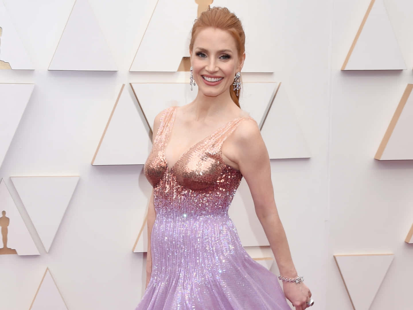 jessica chastain 桌布 壁紙 — 一位微笑的女性身穿閃爍玫瑰到薰衣草色亮片禮服，在白色幾何奧斯卡背景前擺姿勢。