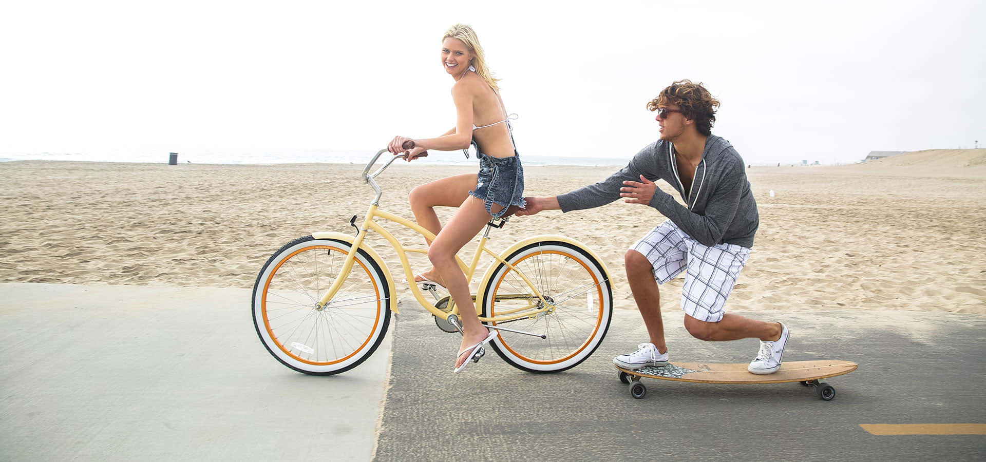 Beach Cruiser 桌布 壁紙 — 一位微笑的女性騎著黃色沙灘巡航自行車，一名踩滑板的男性沿著沙灘旁的鋪砌小徑推著她。