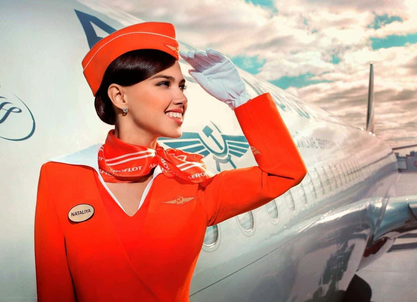 Flight Attendant 桌布 壁紙 — 一位微笑的女空中服務員穿著鮮橙色制服，在陰天下一架白色俄羅斯航空飛機旁敬禮。